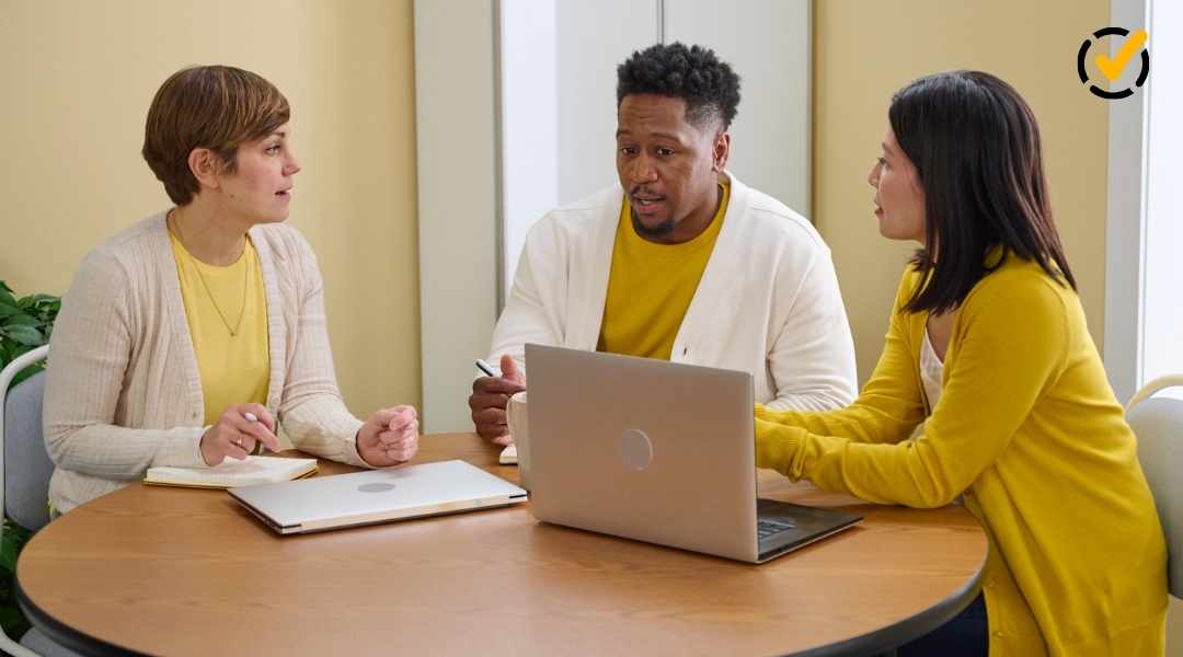 Three diverse teachers—two women and one man—sitting at a round wooden table discussing data on a laptop and in notebooks. The "Balanced Assessment" checkmark logo is subtly placed in the top right.