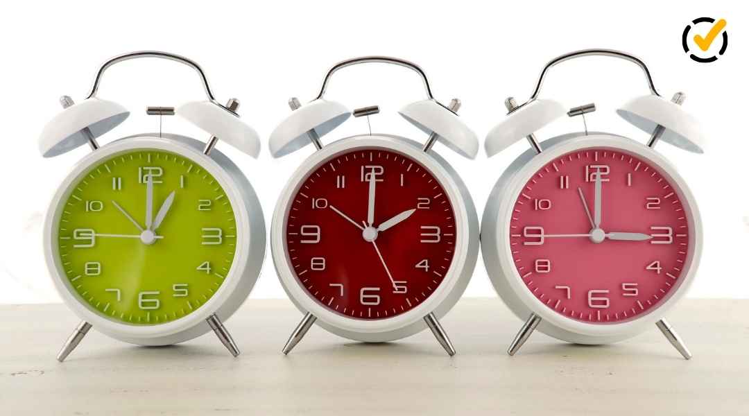 Three colorful alarm clocks (lime green, red, and pink) lined up against a white background, representing time changes for Daylight Saving