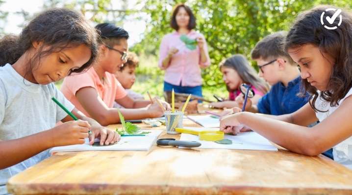 A group of diverse elementary students sitting at an outdoor wooden table, drawing and using magnifying glasses to study leaves with a teacher.