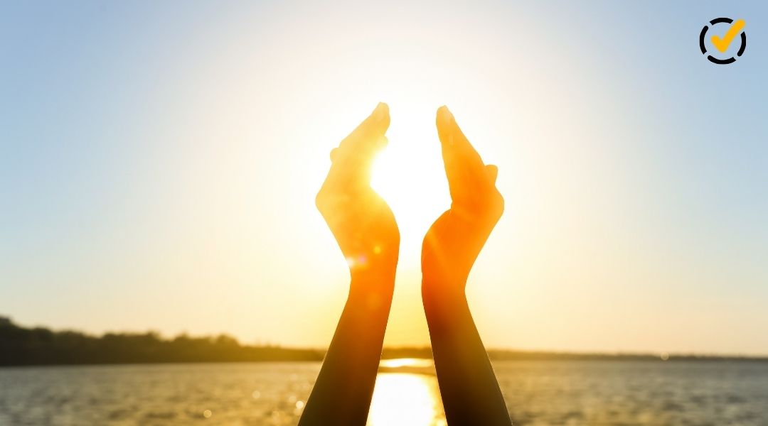 Hands reach toward the sun over a body of water on a bright summer day.