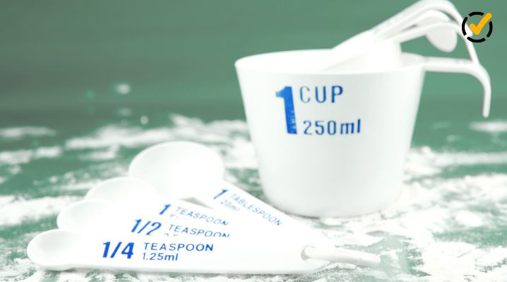 White measuring cup and measuring spoons labeled with cup, tablespoon, and teaspoon measurements on a flour-covered surface.