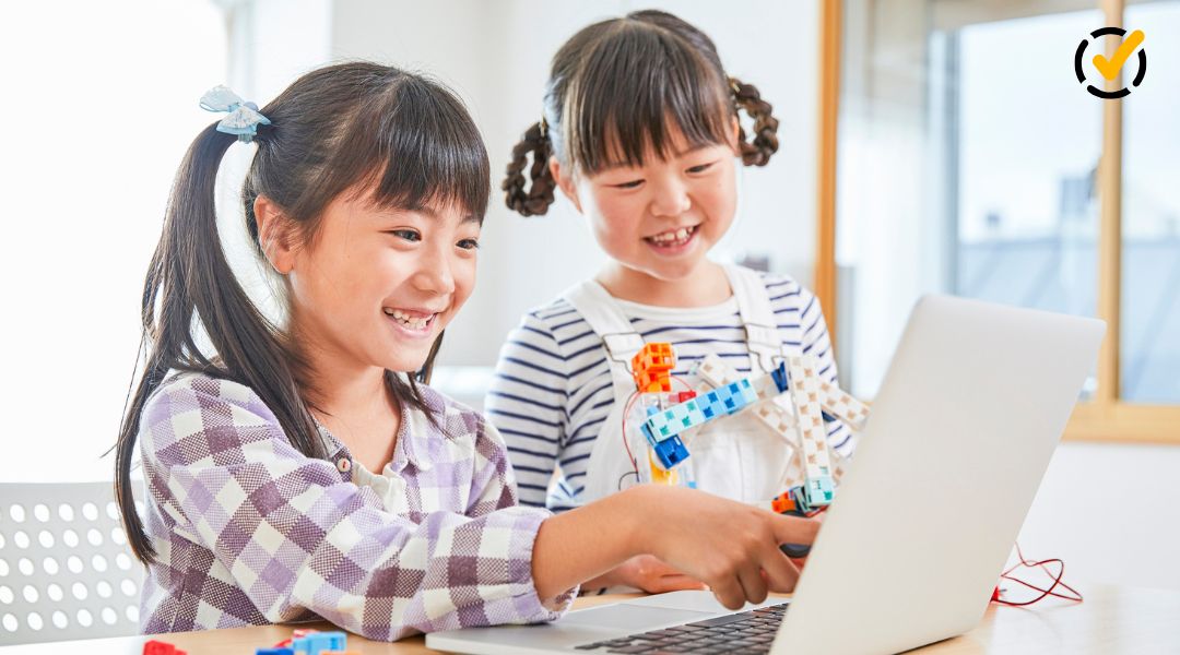 Two elementary students use a laptop and a colorful robotics model during a hands-on STEM activity involving technology and engineering.