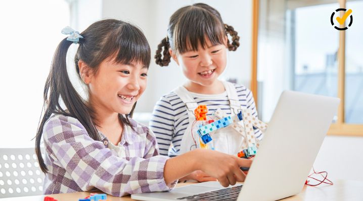 Two elementary students use a laptop and a colorful robotics model during a hands-on STEM activity involving technology and engineering.