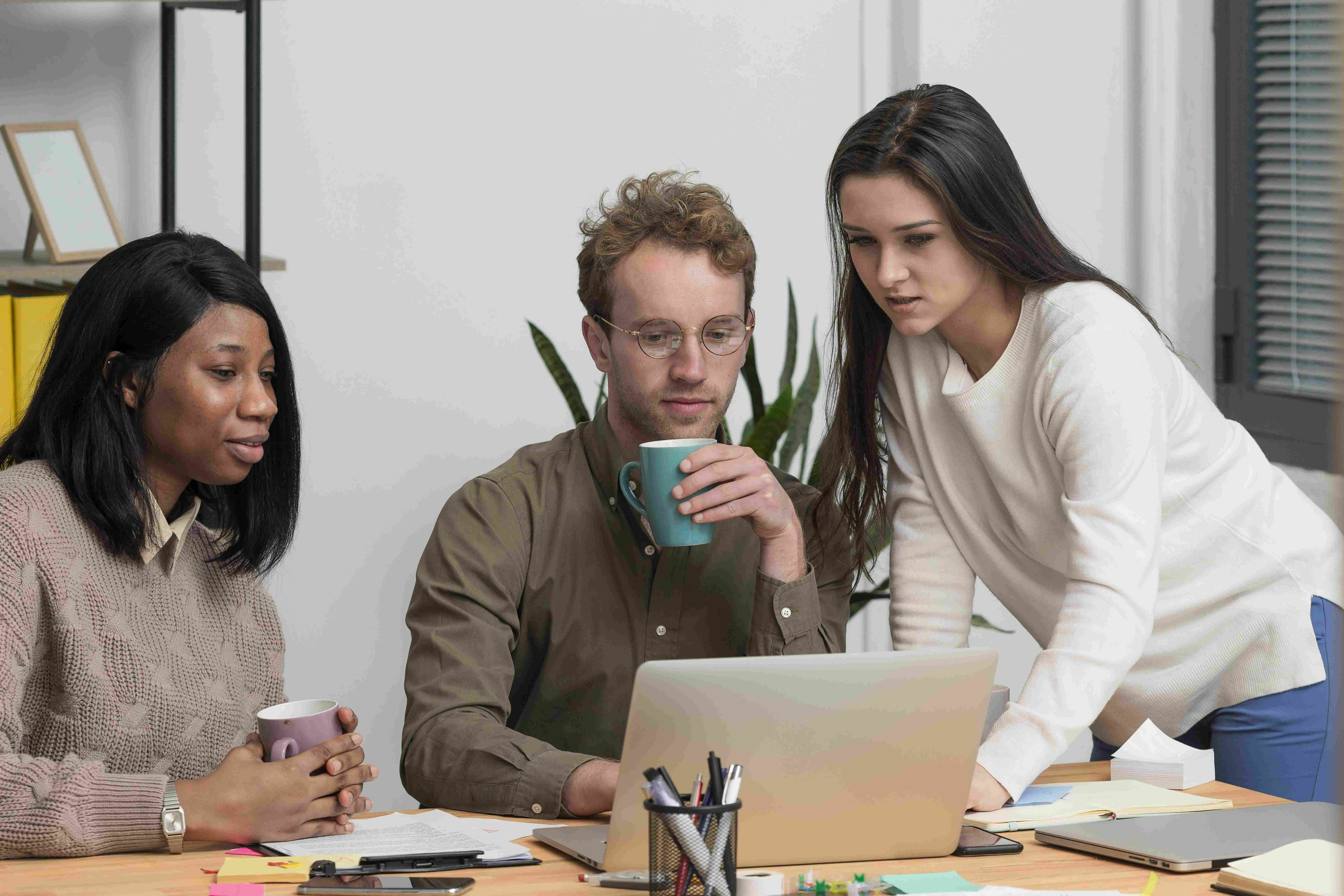 Um homem e duas mulheres, segurando canecas, ao redor de um notebook, trabalhando.