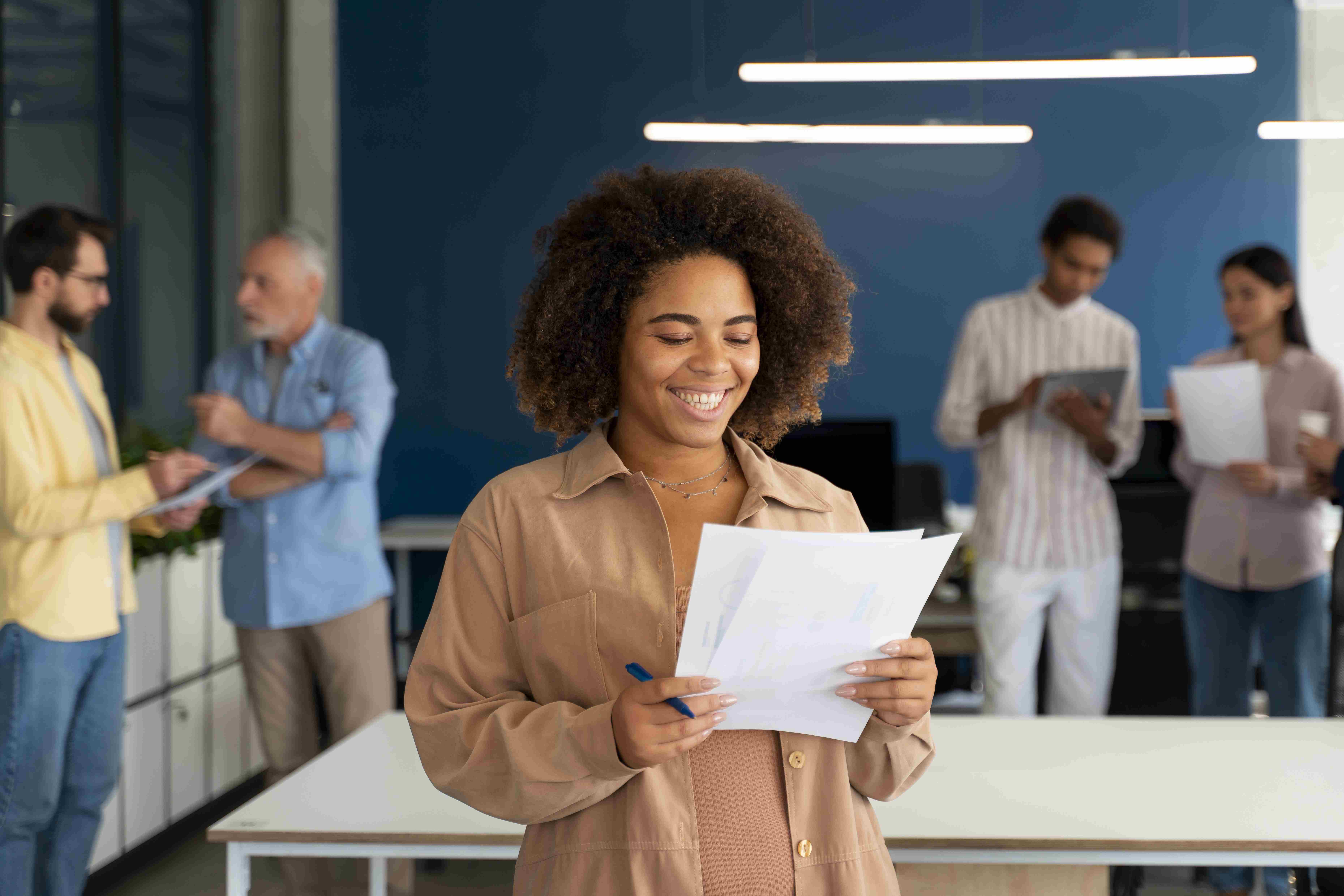 Mulher à frente, segurando papéis, com colegas de trabalho conversando ao fundo, em escritório da empresa.