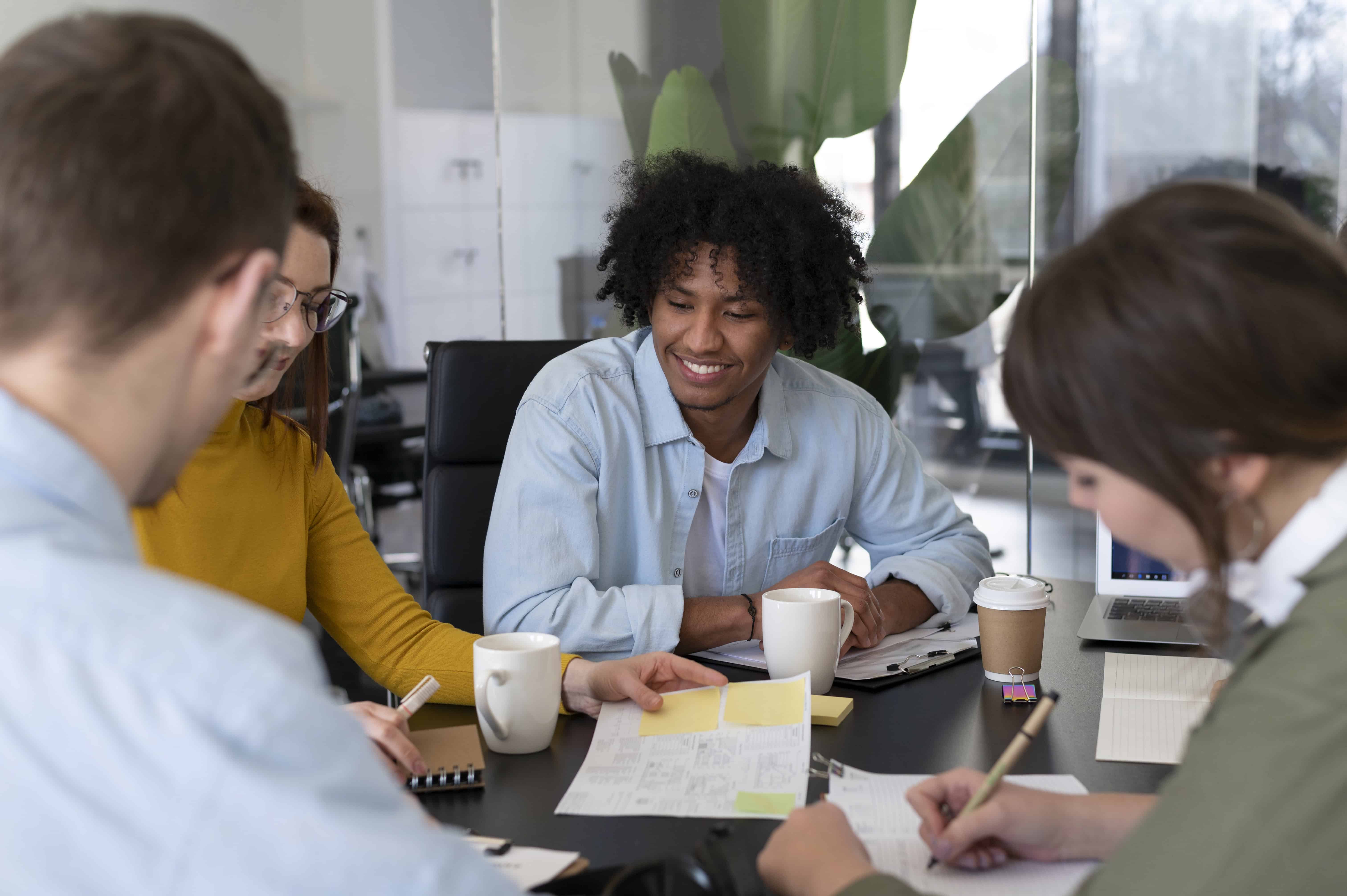 Time de trabalho reunido ao redor da mesa, com papéis e copos de café, conversando.