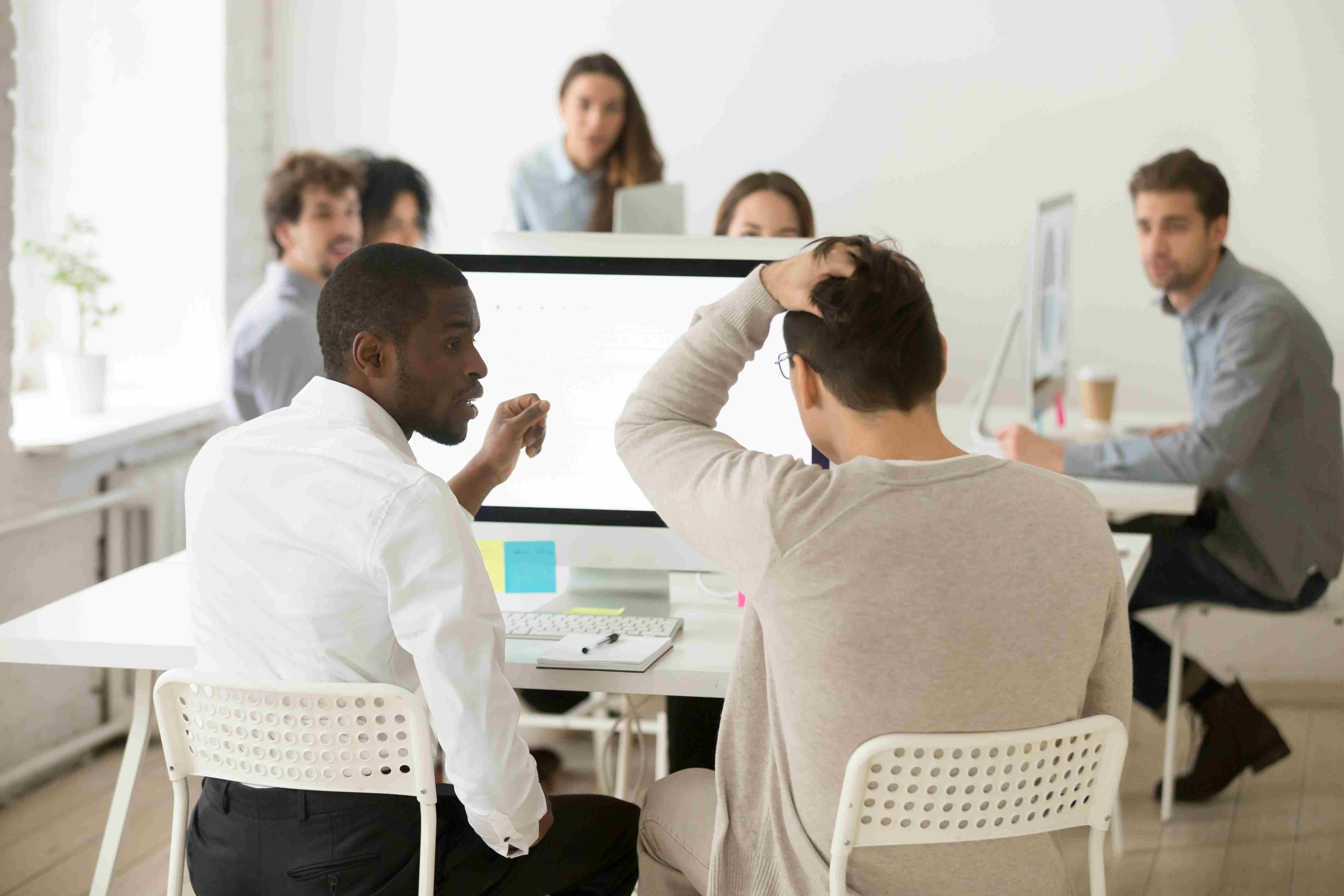 Dois homens sentados no computador. Um deles está com a mão na cabeça, apreensivo. Ao fundo, está o restante da equipe.