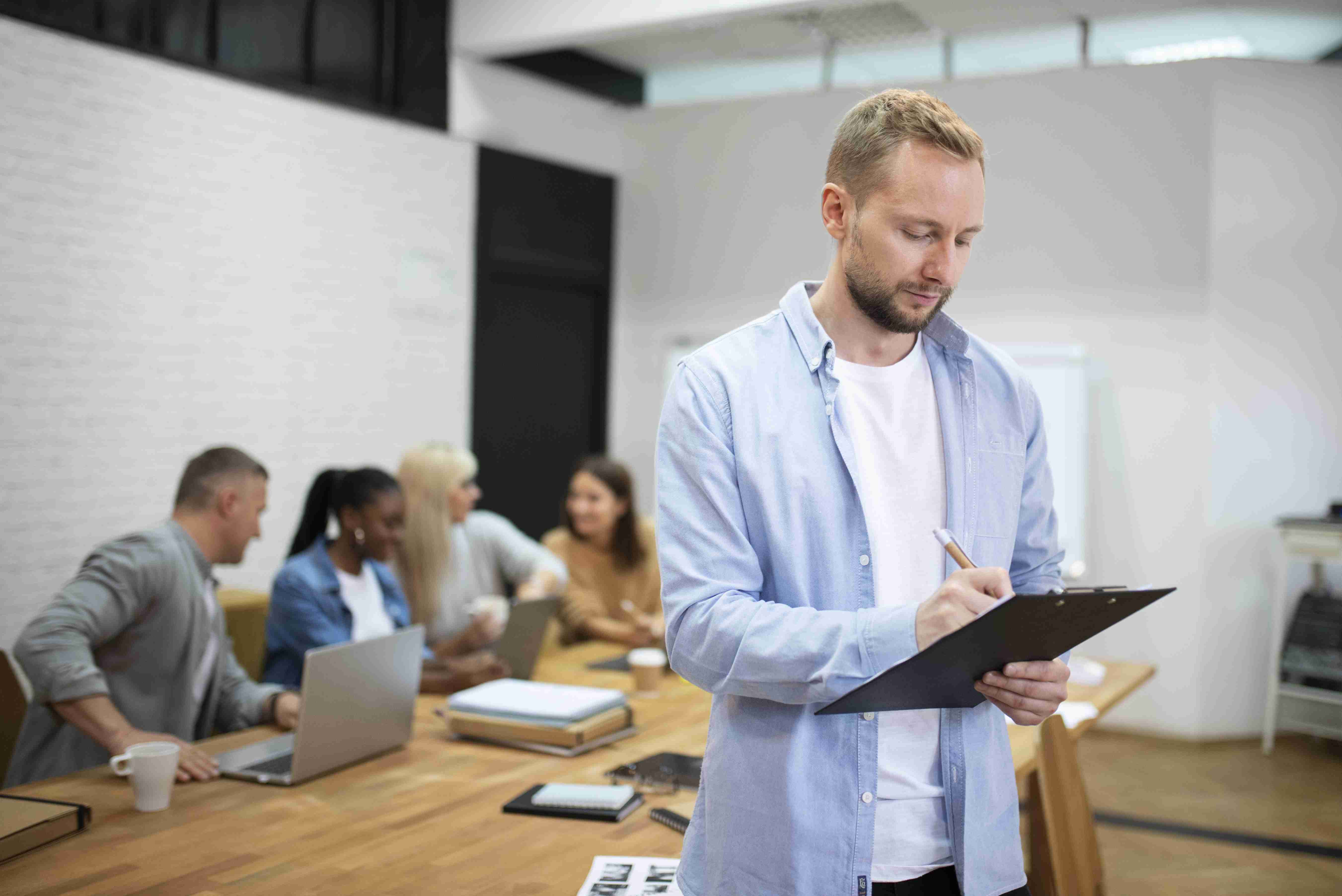 Homem de camiseta branca e camisa azul por cima, tomando notas. Ao fundo, equipe de trabalho conversando.