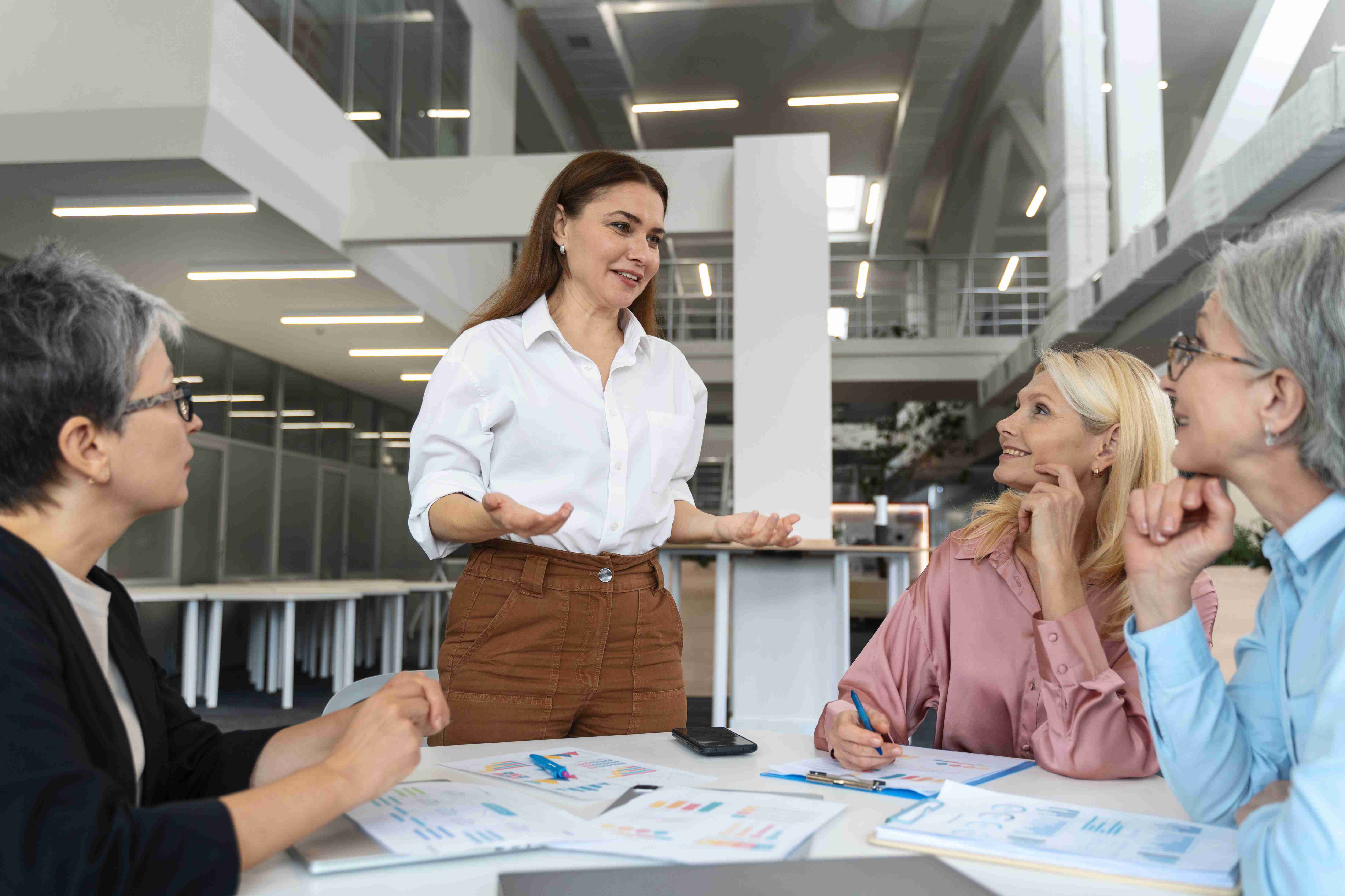 Liderança técnica realizando treinamento para três outras mulheres, sentadas à mesa, com papéis ao redor.