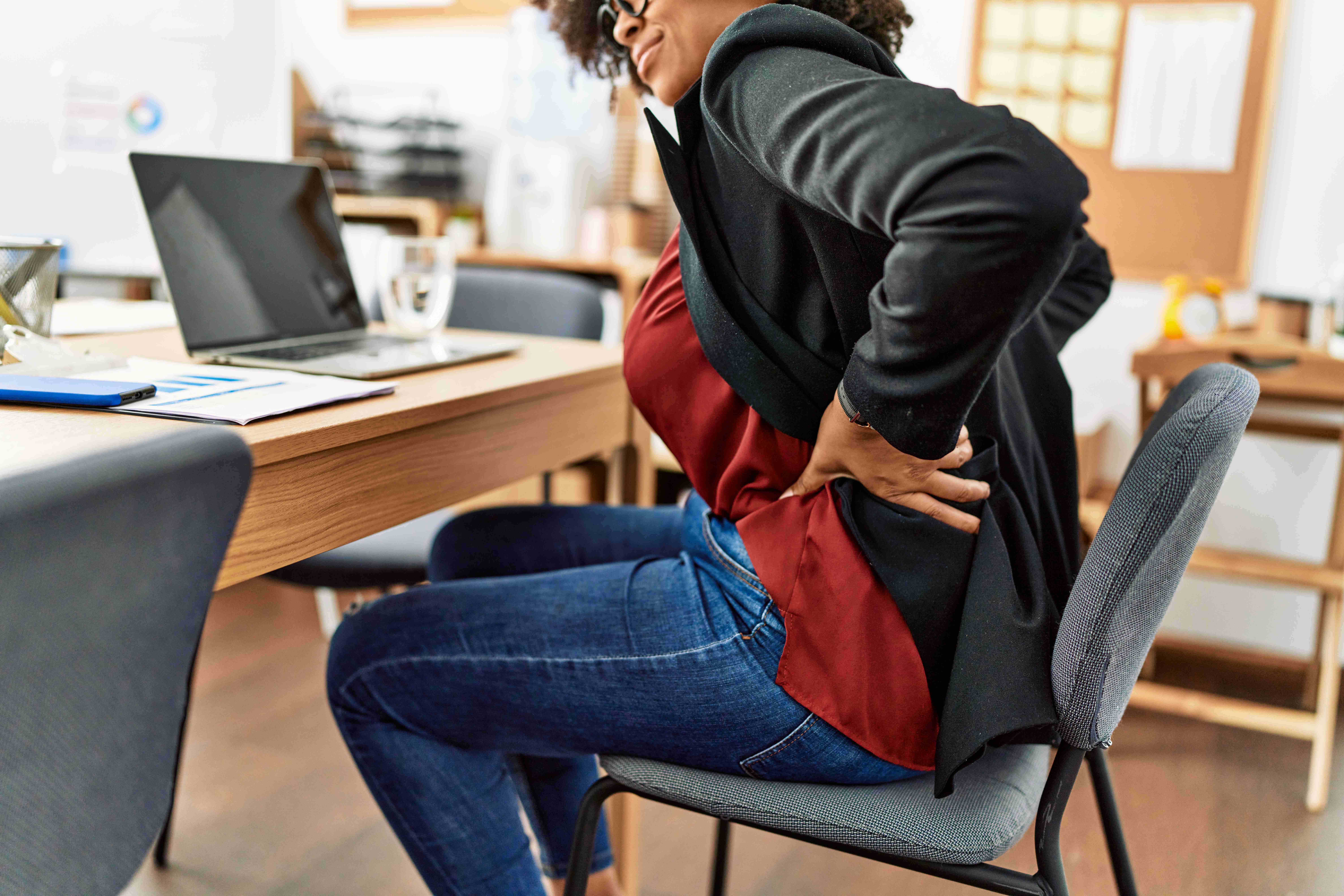 Mulher sentada na cadeira, em frente ao computador, com as mãos na lombar e expressão de dor.