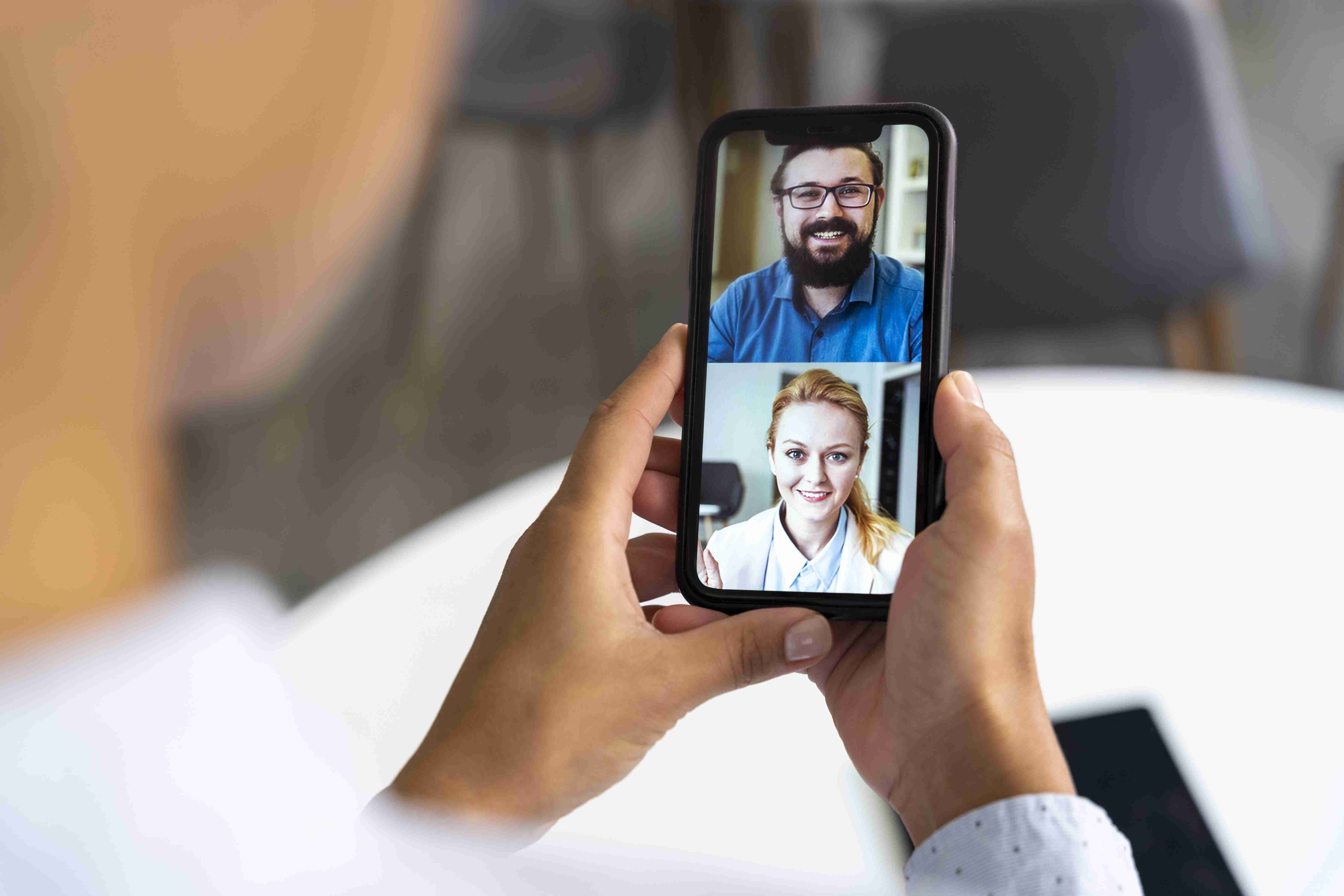 Pessoa segurando celular. Na tela, um homem de barba e uma mulher loira, ambos sorrindo, enquanto fazem reunião.