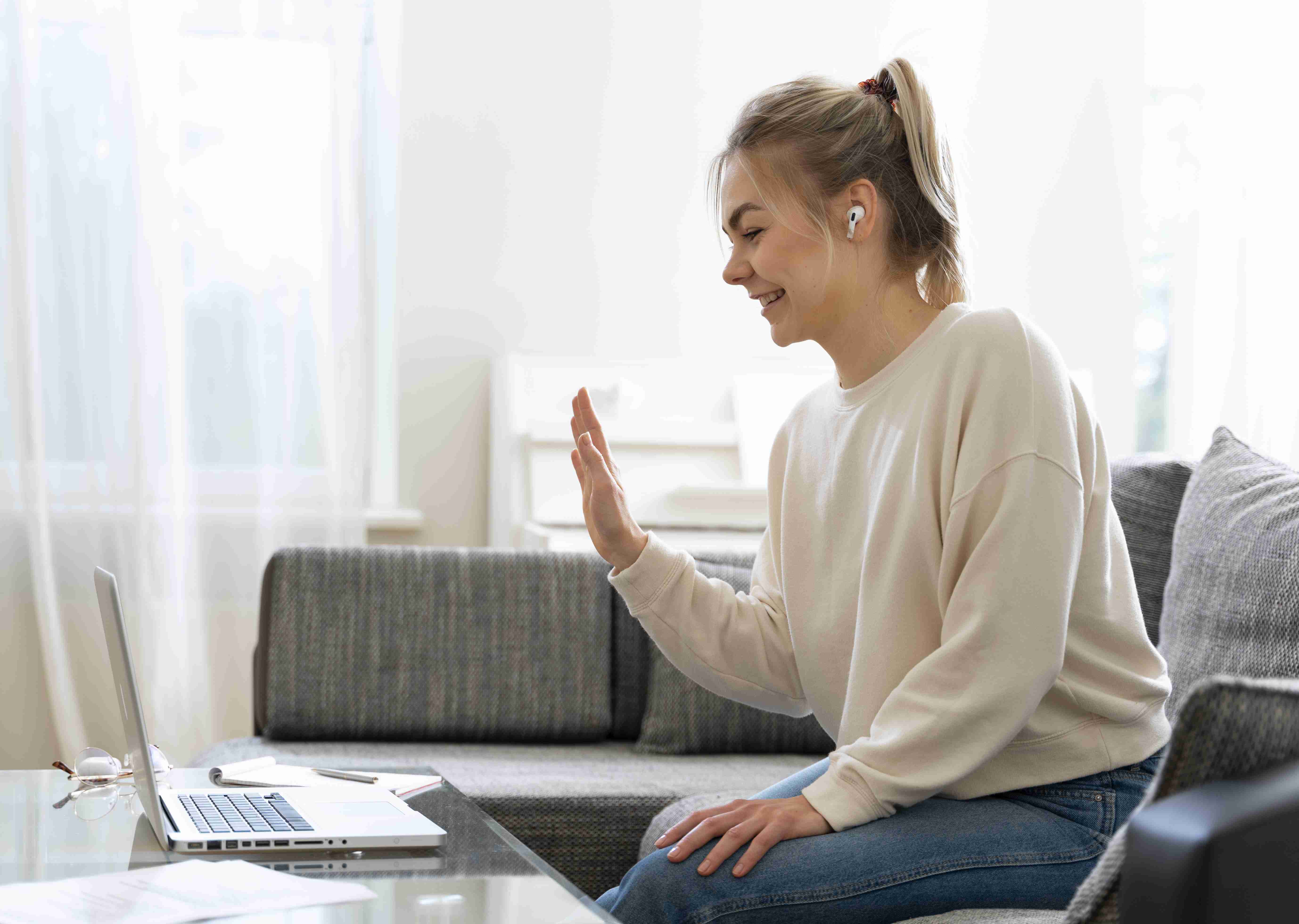 Mulher loira de moletom, sentada em um sofá, acenando para a tela do notebook, durante consulta online.