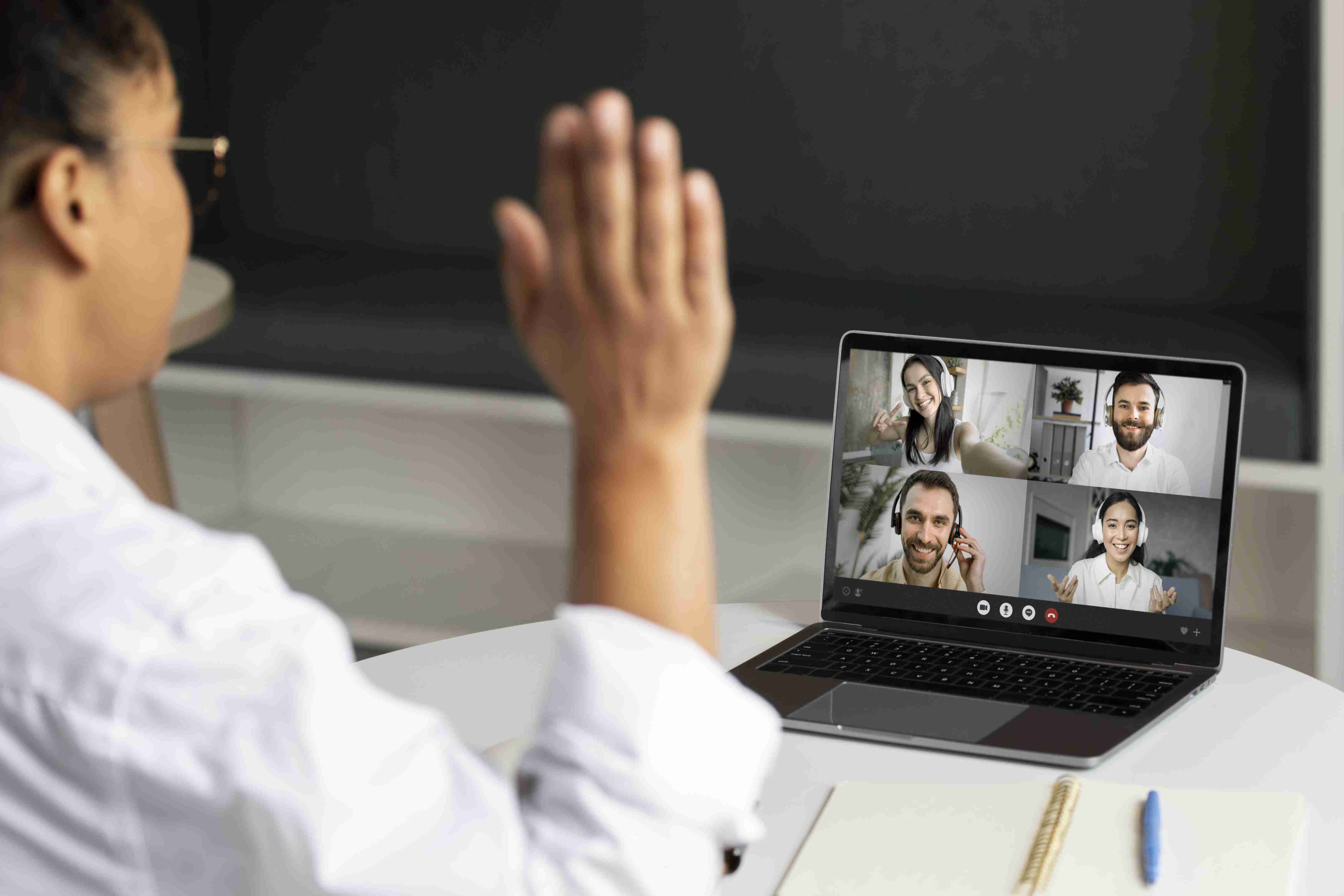 Mulher acenando para quatro colegas de equipe na tela de um notebook, durante reunião de coordenação do cuidado.