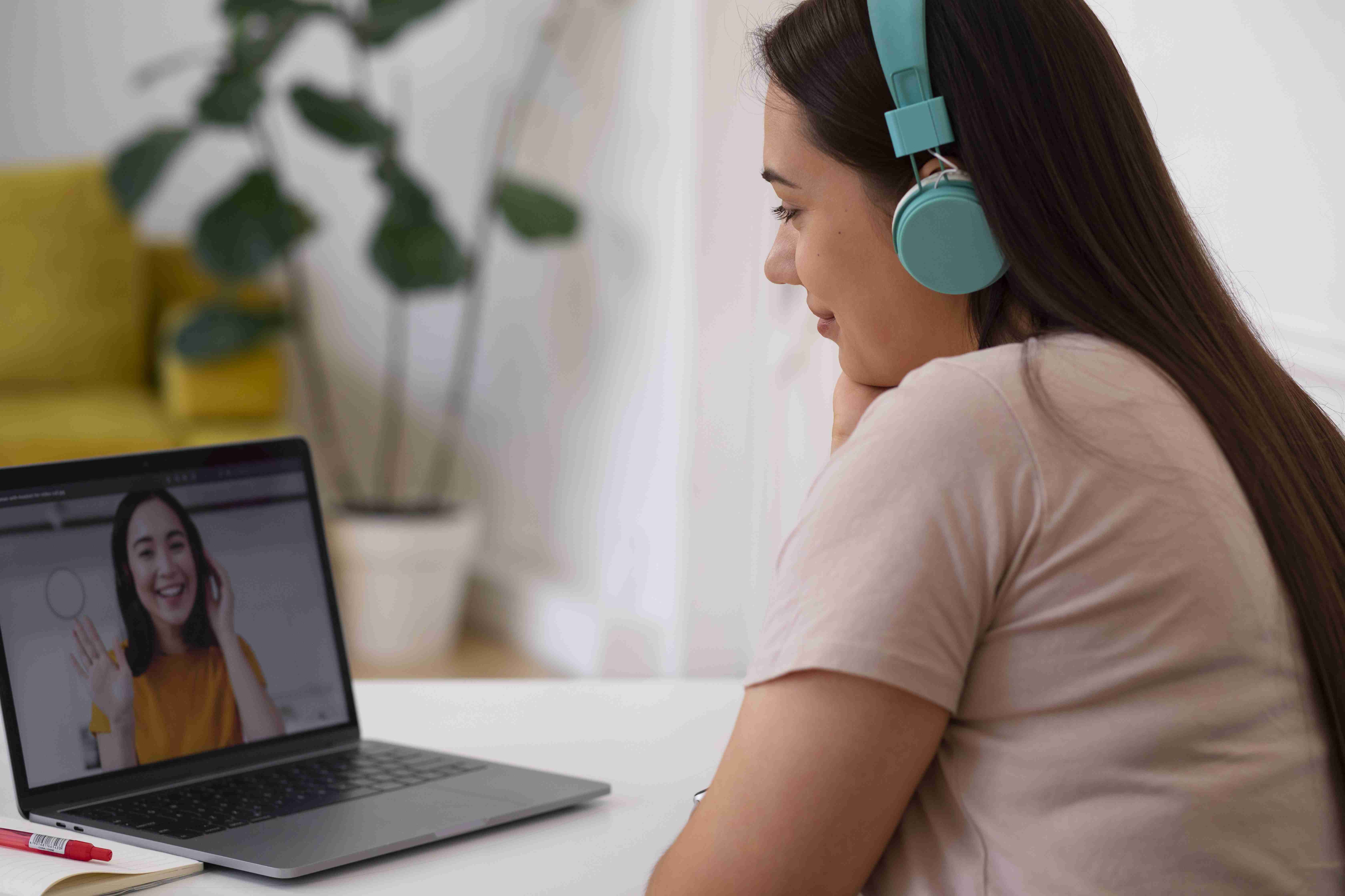 Mulher de fones de ouvido e camiseta rosa bebê, realizando consulta virtual com médica, que sorri na imagem do notebook.