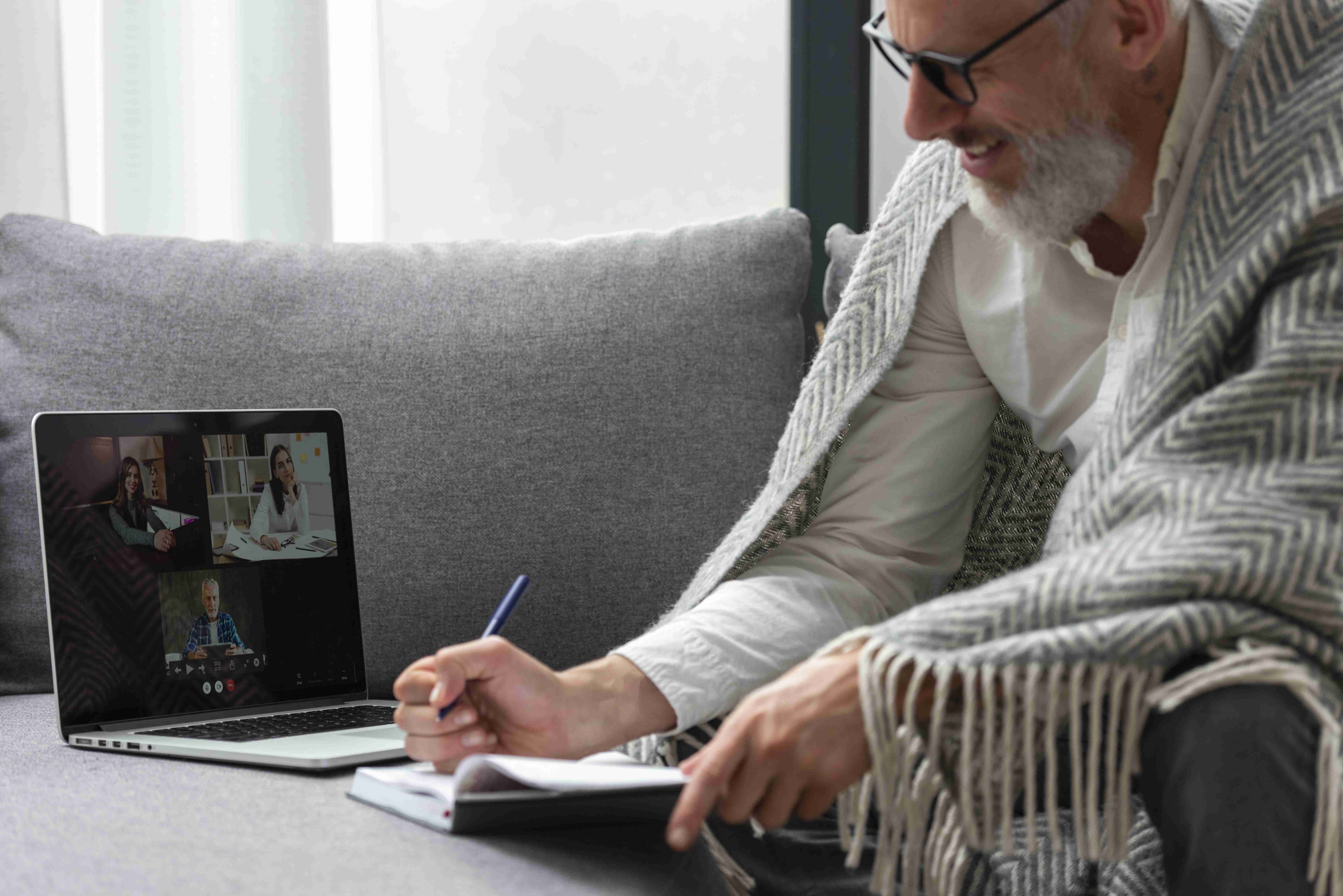 Homem sentado no sofá, realizando teleconsulta com equipe de saúde integrativa, anotando em um caderno.