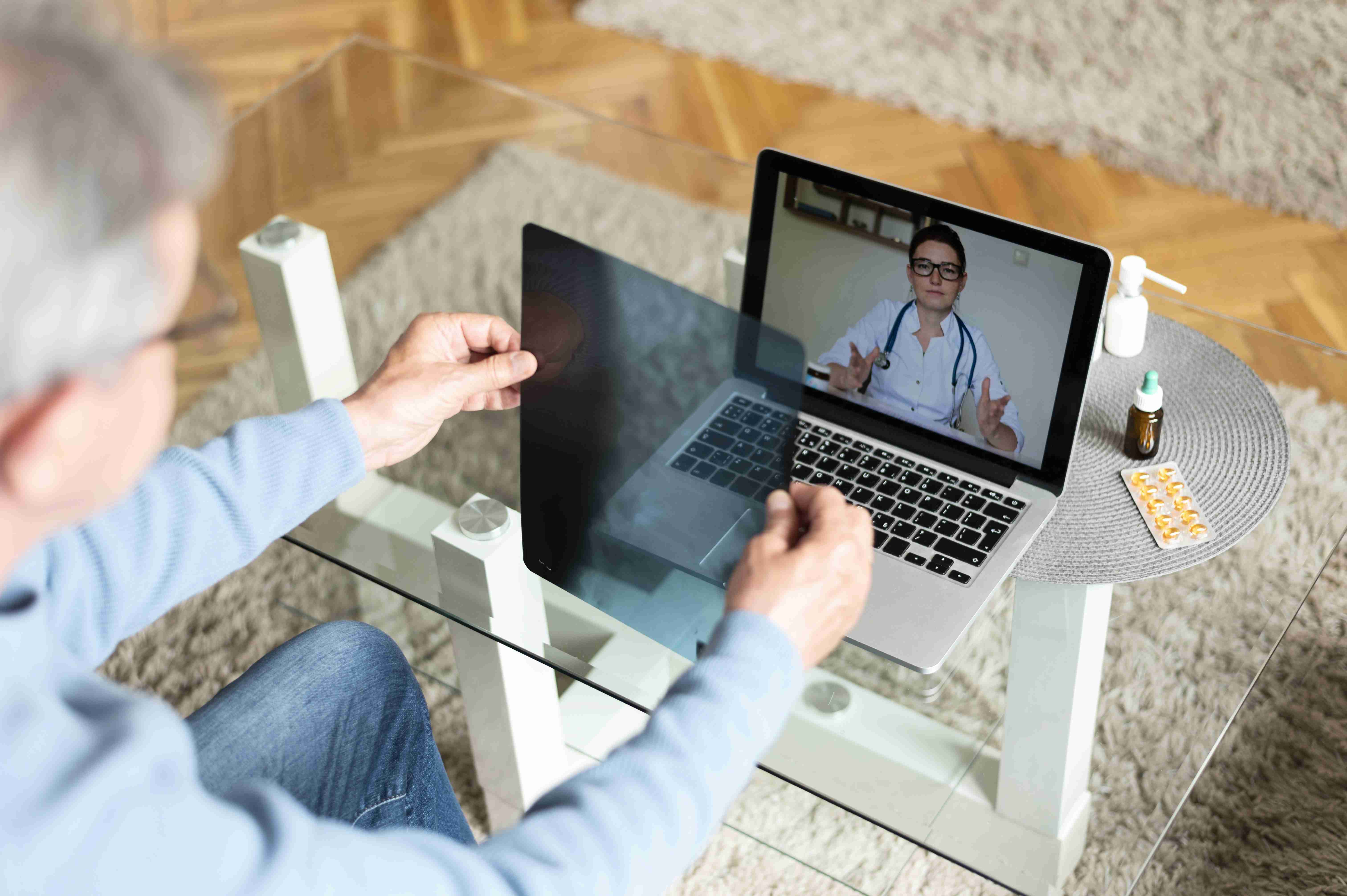 Homem mostrando raio-x para médico, enquanto realiza consulta virtual sentado no sofá.