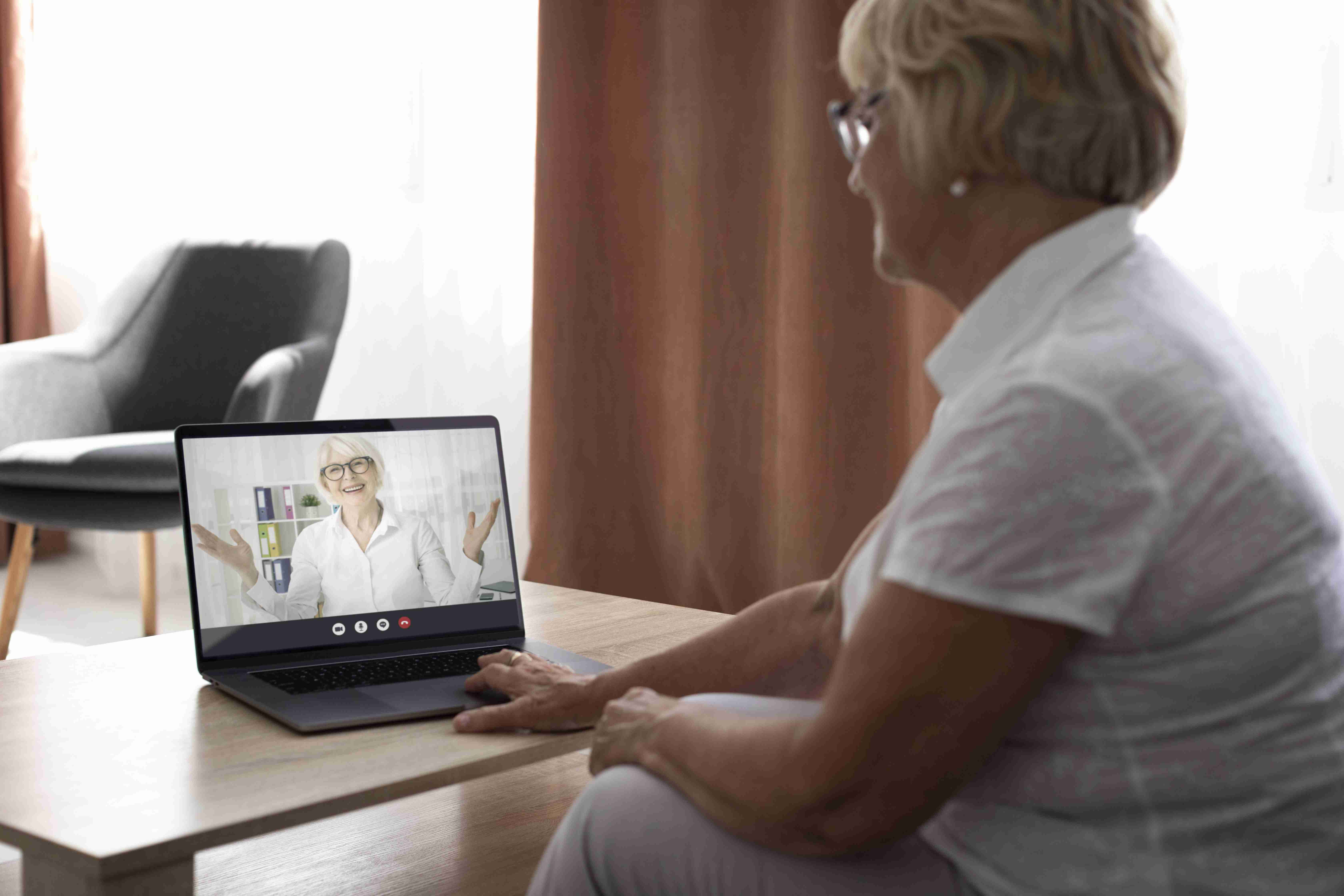 Senhora sentada no sofá, em uma sala com móveis de madeira, realizando teleconsulta pelo notebook com médica.