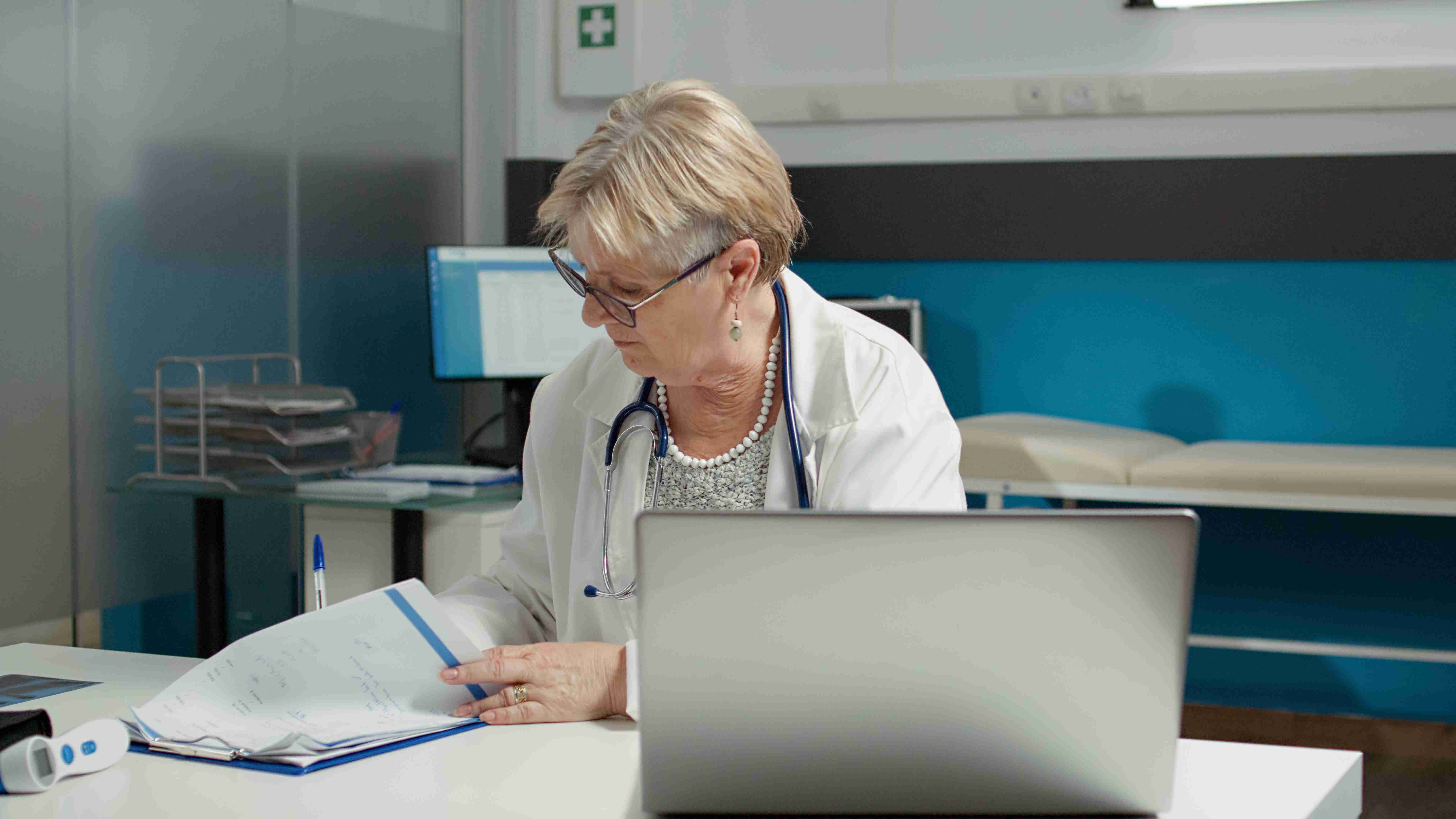 Médica escrevendo na ficha do paciente em frente ao notebook, enquanto digitaliza informações sobre o paciente.