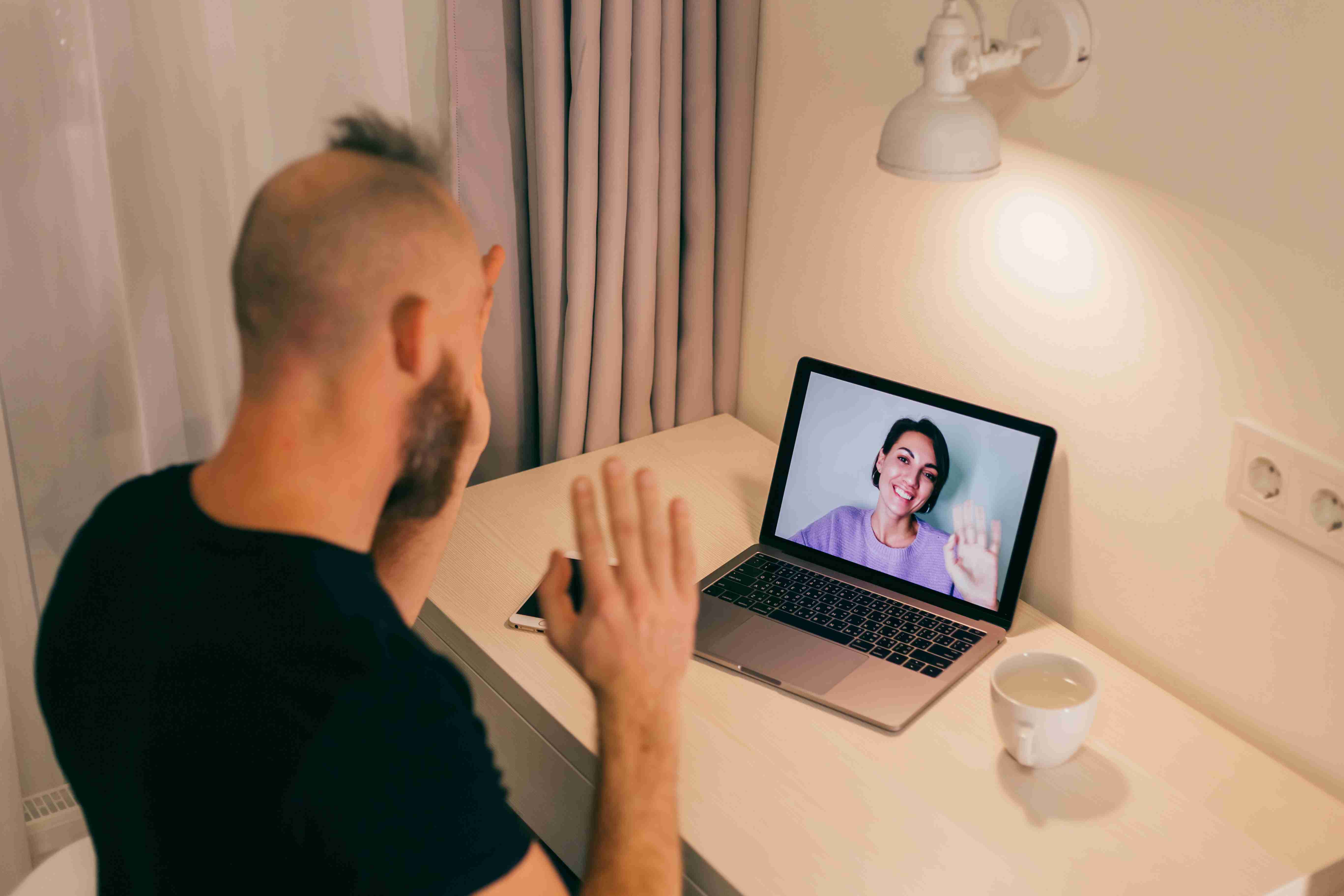 Homem realizando teleconsulta. No notebook, uma profissional de saúde sorri e acena para ele.