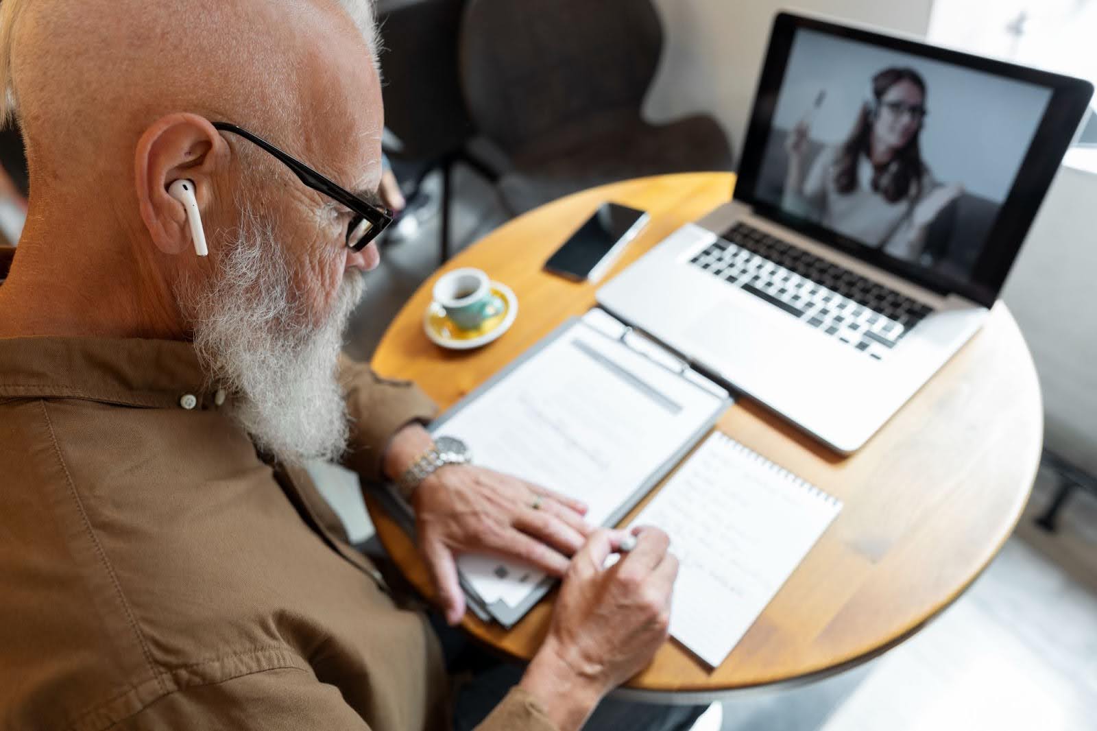 Homem idosos fazendo anotações em um caderno durante teleconsulta com médica.