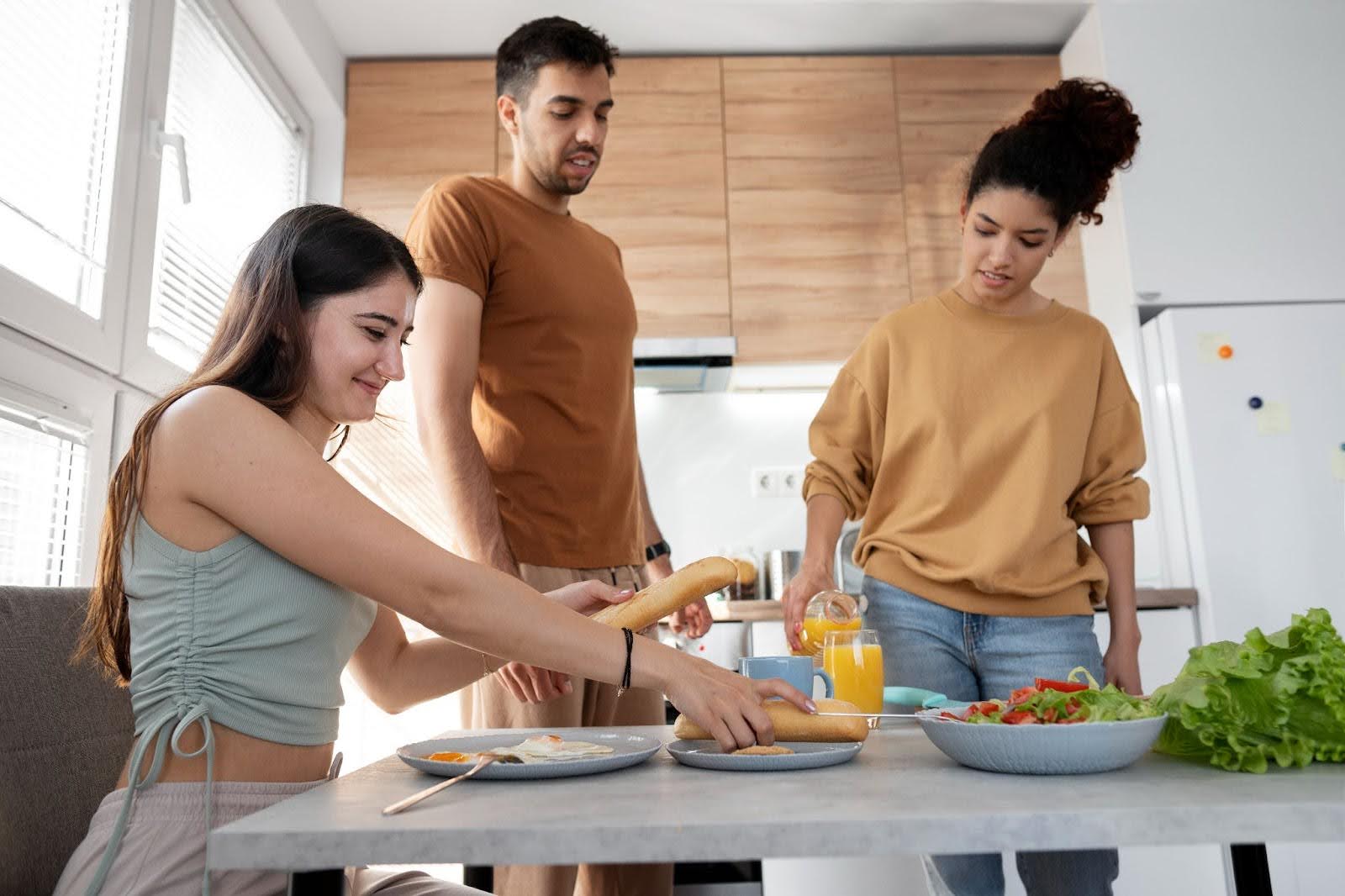 Uma mulher sentada à mesa, com um pão na mão, e dois amigos em pé, ao lado dela, conversando e servindo suco de laranja.
