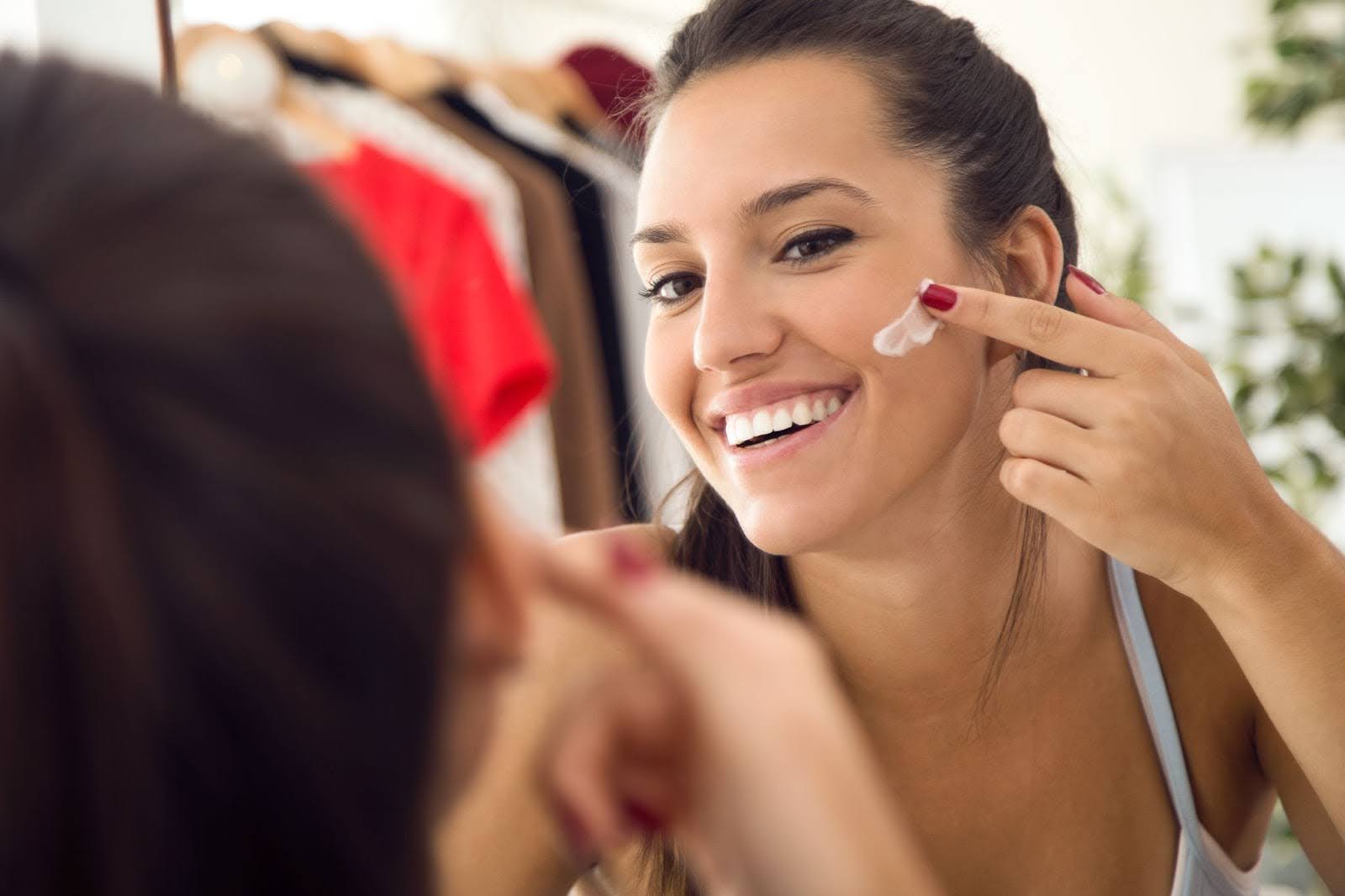 Mulher no espelho, sorrindo enquanto passa protetor solar no rosto.