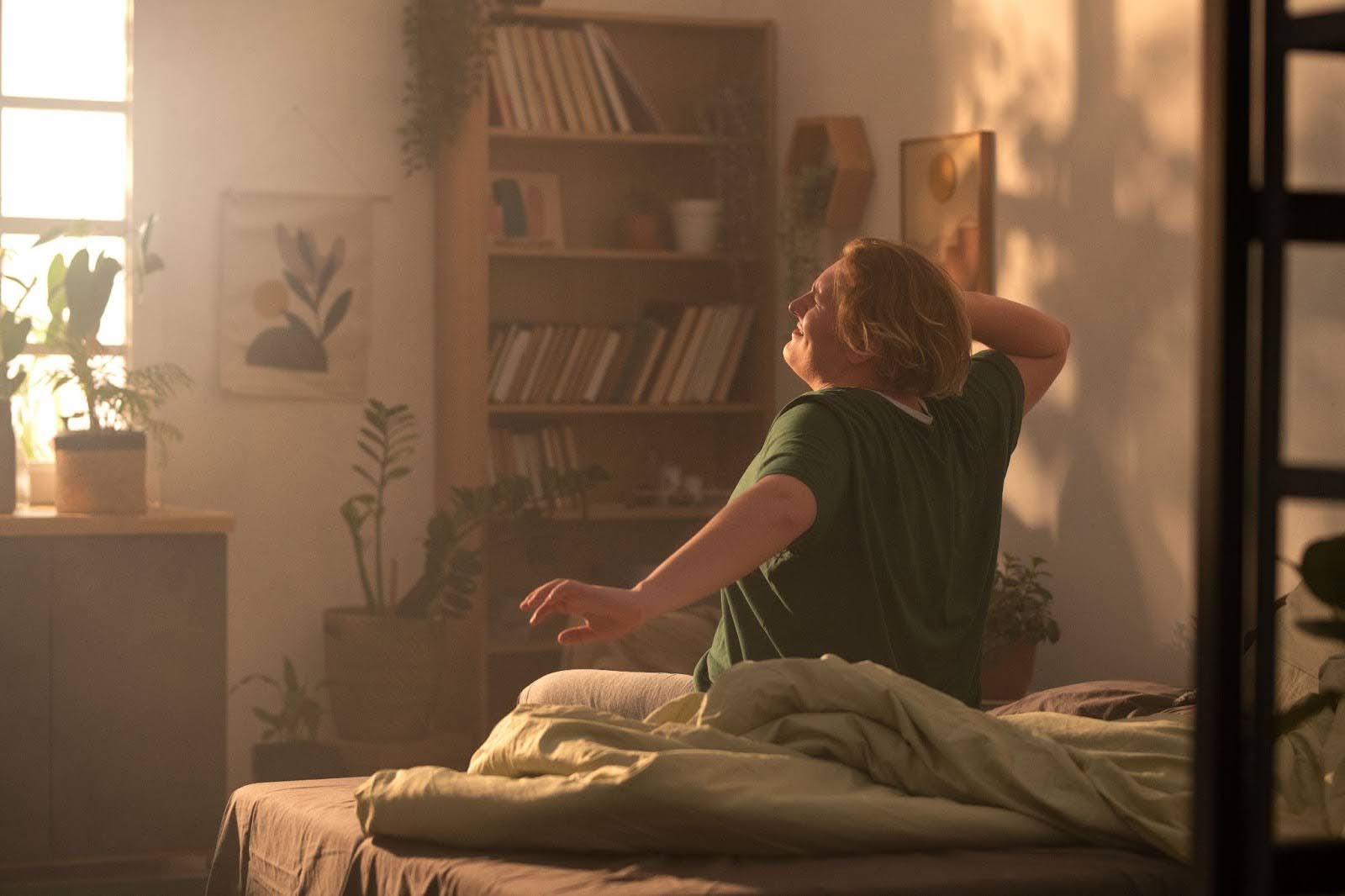 Pessoa se espreguiçando na cama, logo ao acordar, com luz do sol entrando pela janela.