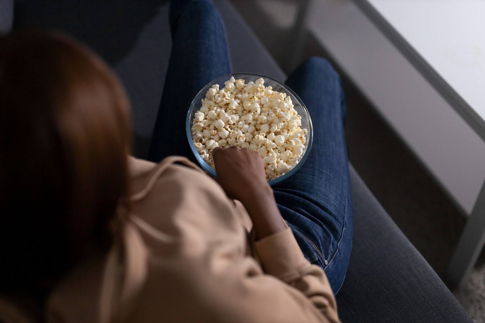 Foto panorâmica de uma mulher sentada no sofá, com um balde de pipoca entre as pernas.