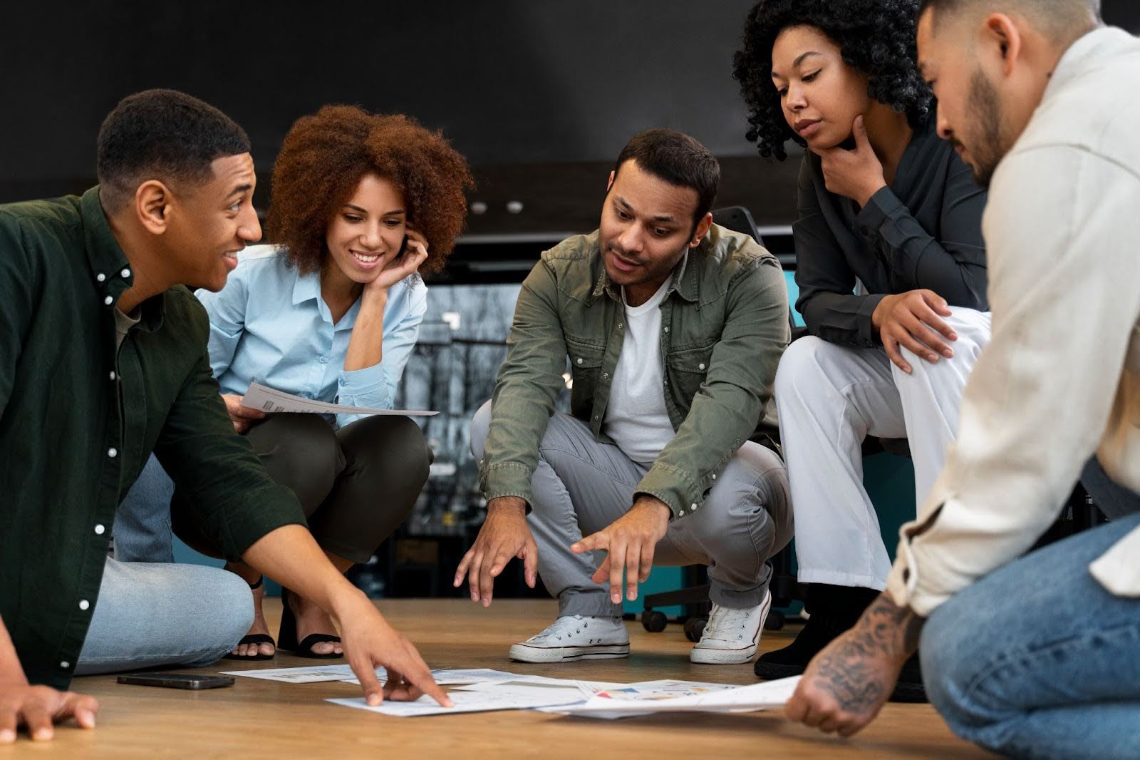 Equipe de trabalho composta de 3 homens e 2 mulheres sentados no chão, ao redor de papeis, durante dinâmica.