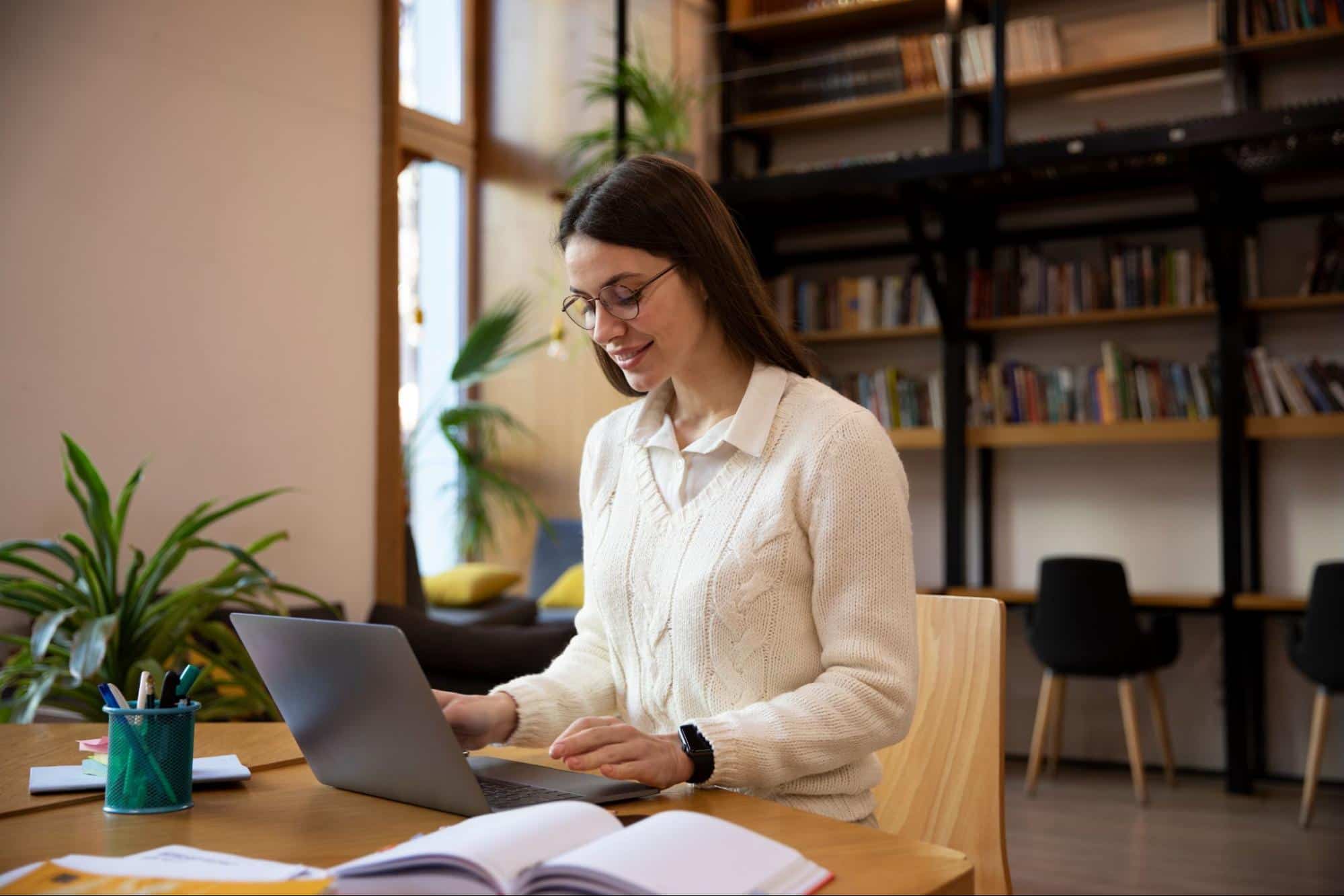 Mulher sentada em frente ao notebook, em um espaço amplo, com estantes de livros e cadeiras.