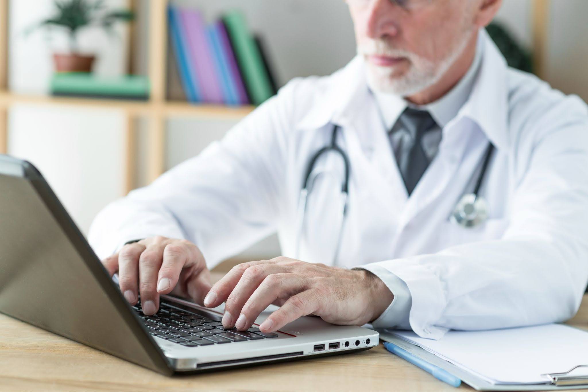 Médico sentado à mesa, digitando em um notebook.
