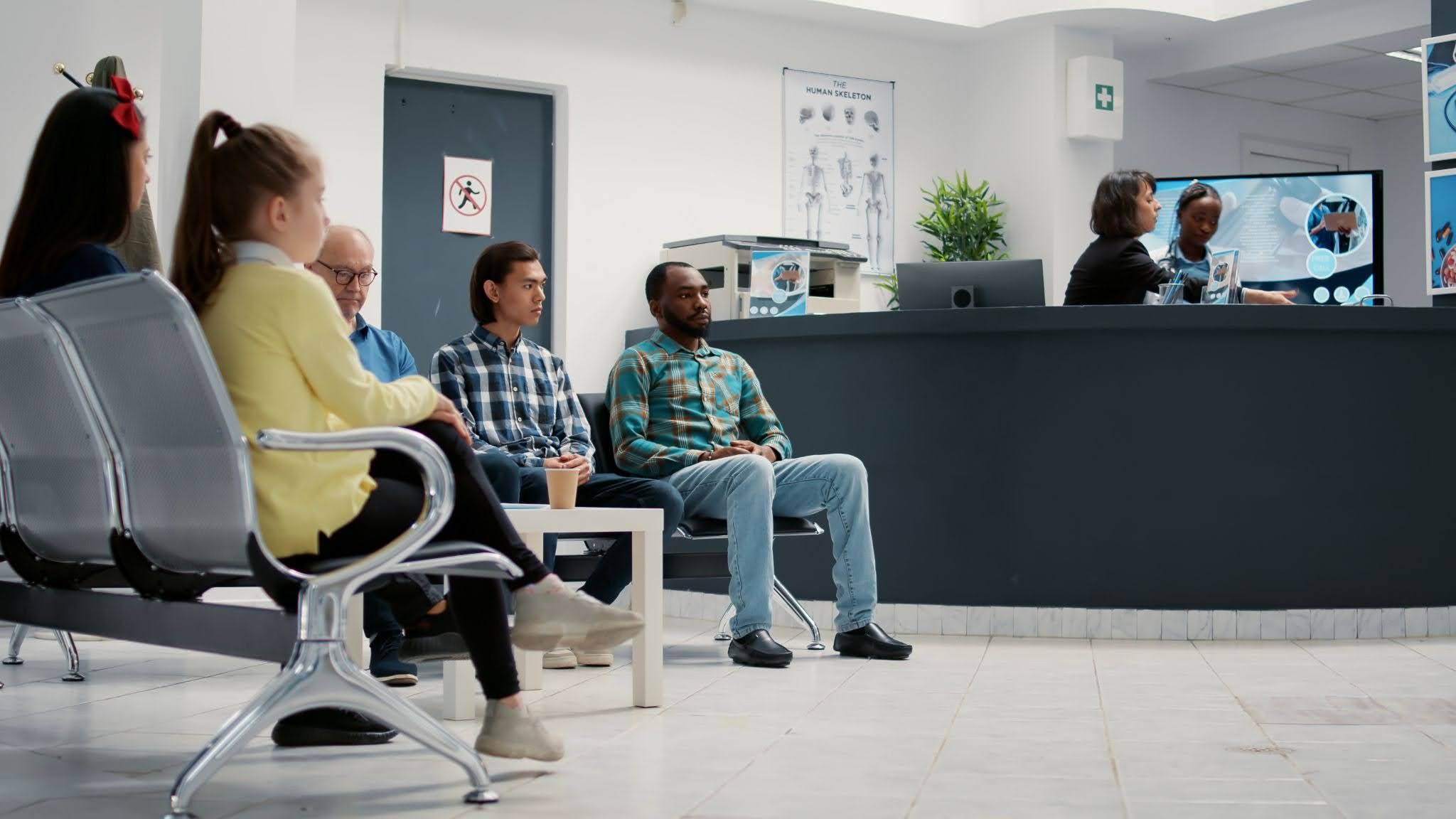 Pessoas em uma sala de espera do hospital, sentadas, com profissionais realizando o atendimento no fundo.