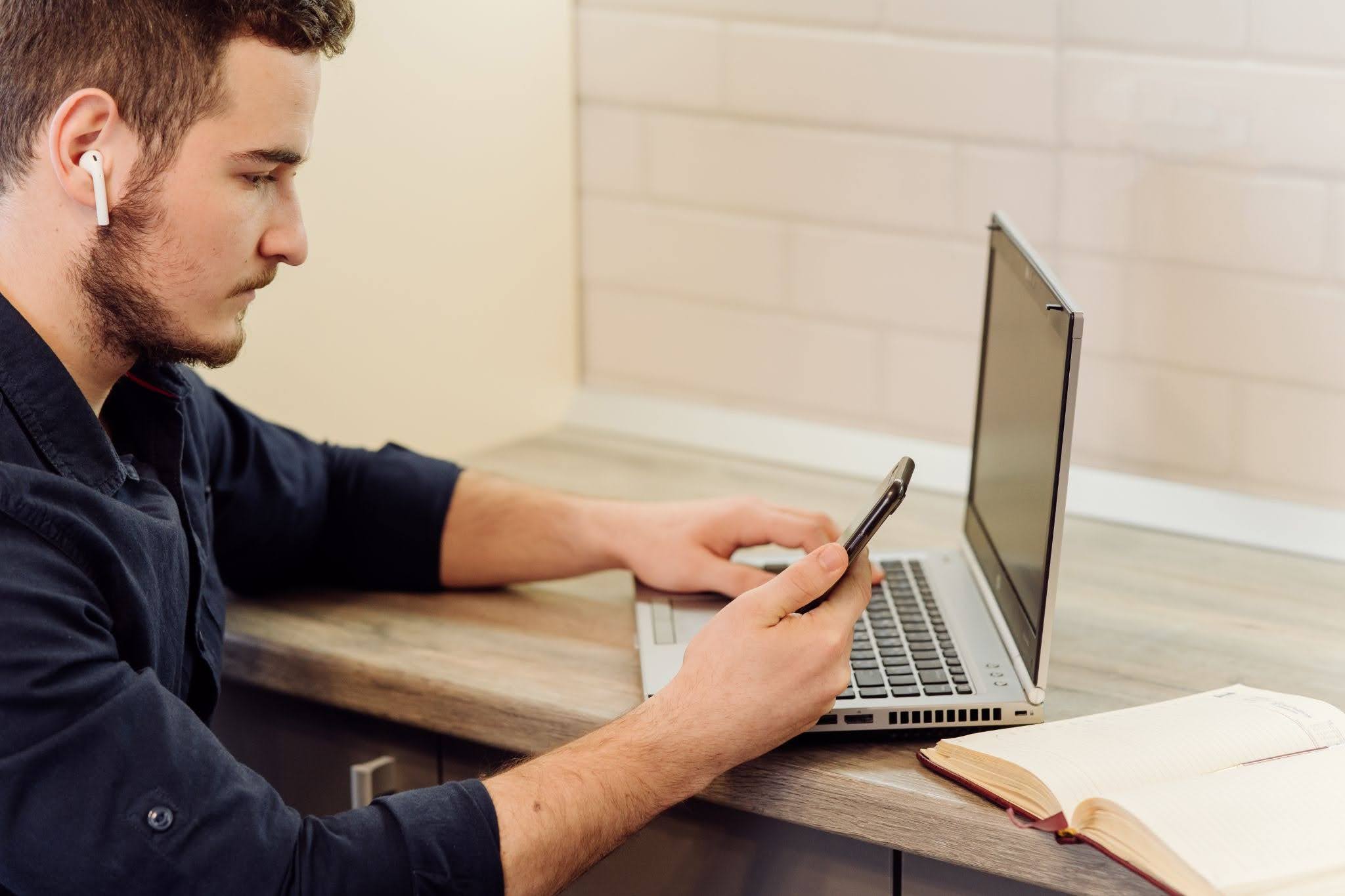 Homem de camisa azul marinho sentado em frente ao notebook, enviando mensagem pelo celular.