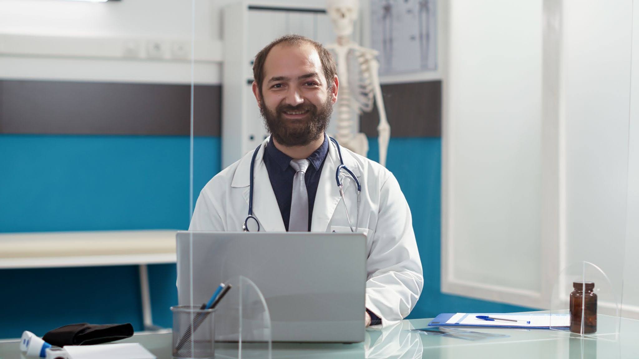 Médico sentado em frente ao notebook, sorrindo enquanto olha para a foto.