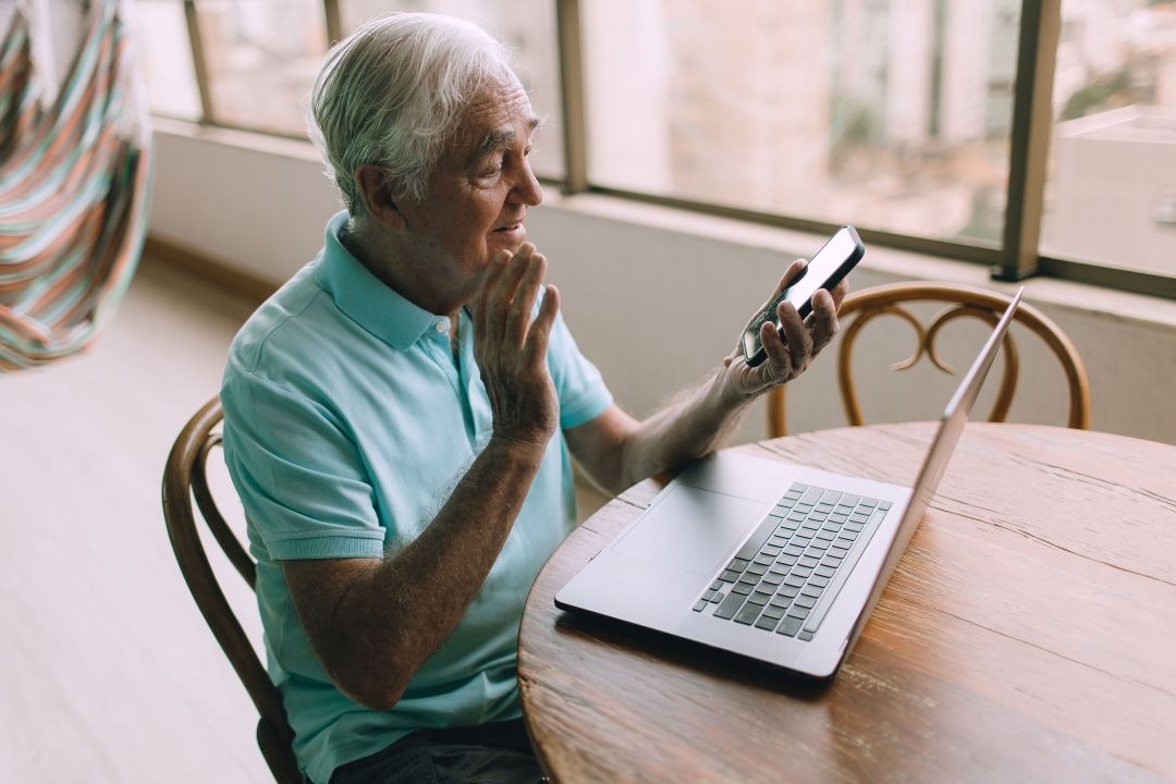  homem idoso segurando celular em frente ao notebook conversando por videochamada