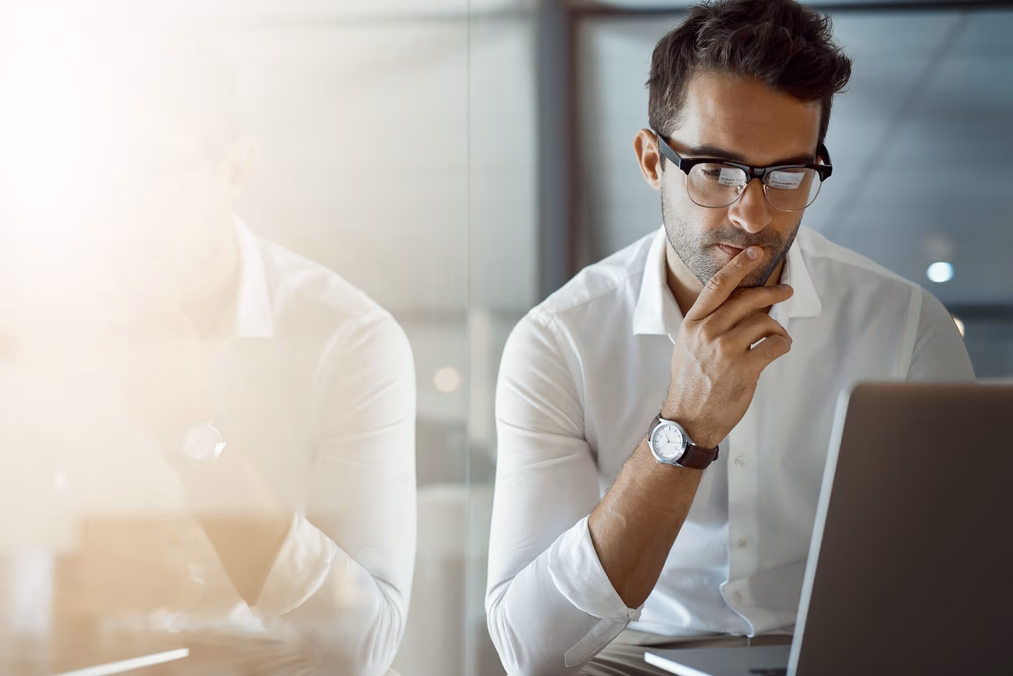 A man in a business setting sits in front of his laptop and thinks about something.