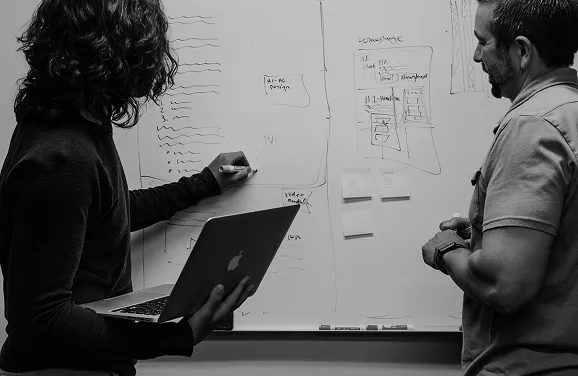 Two people discussing ideas and sketching on a whiteboard, one holding a laptop and the other writing with a marker.