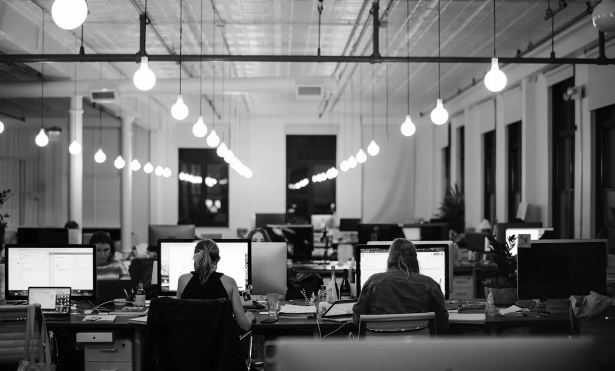 Black and white photo of a modern office with rows of desks, computers, and hanging light bulbs, showing two people working at their desks.