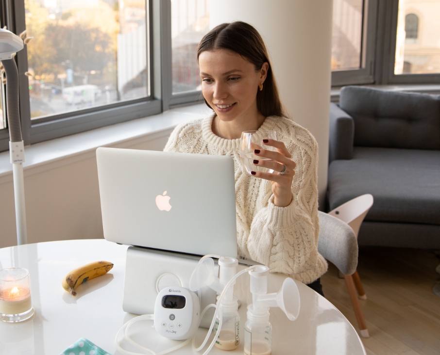 Woman smiling while working on a laptop at a table with a breast pump, in a bright room with large windows.