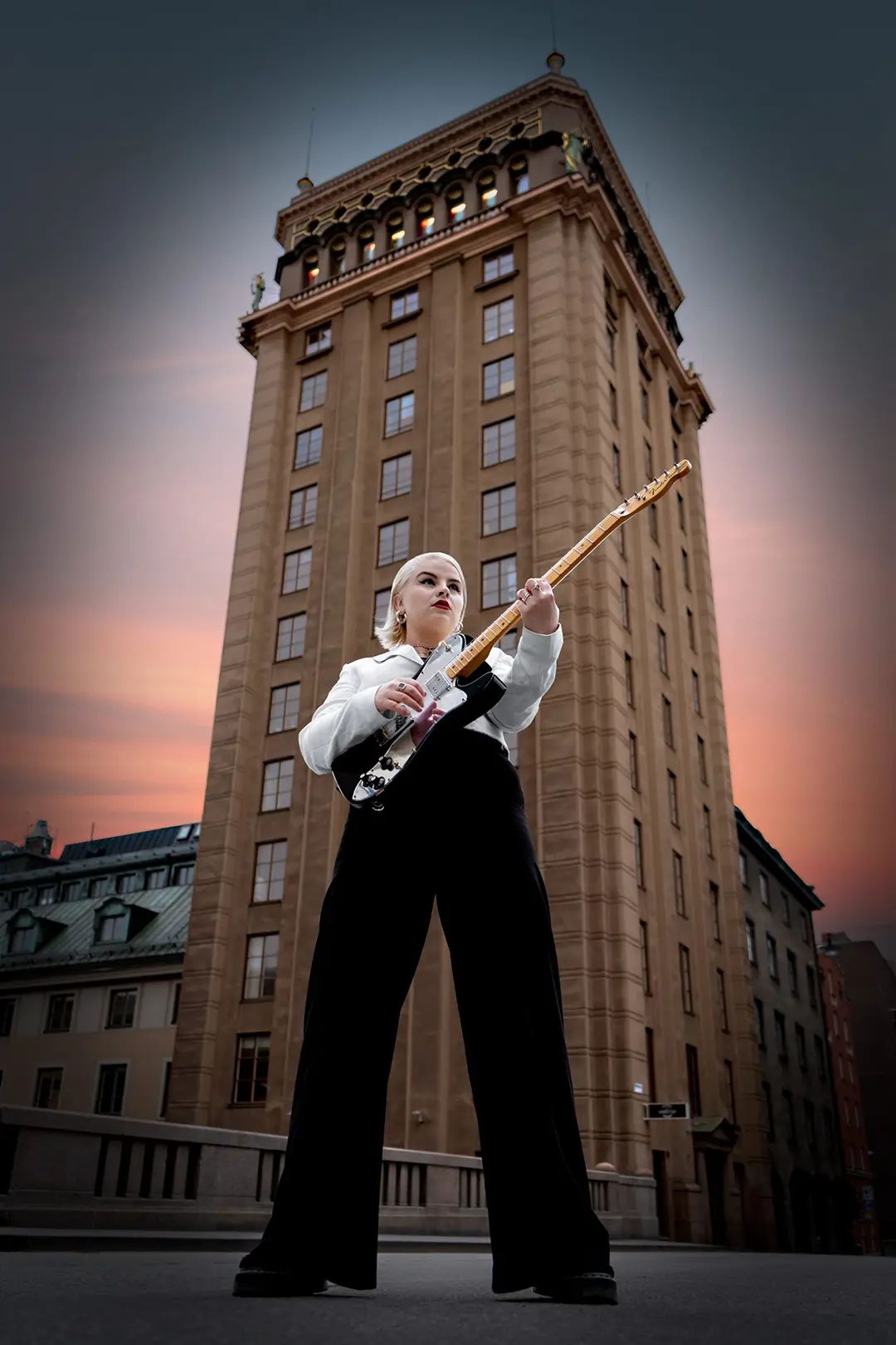Person med blond kort hår spelar elgitarr framför en hög byggnad under solnedgången.