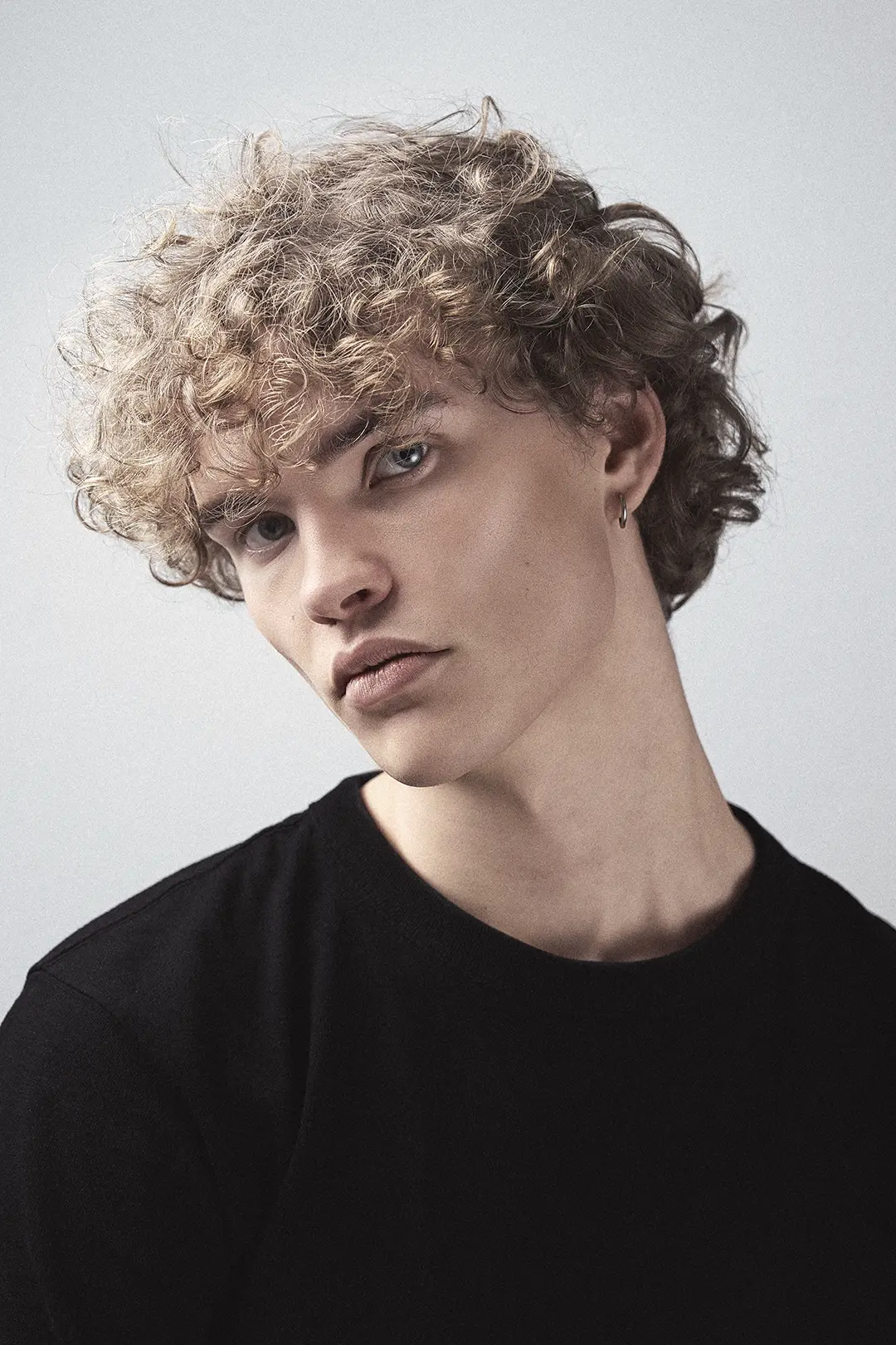 Porträtt av en ung person med lockigt hår, ljus hy och en örhänge i vänster öra, klädd i svart tröja mot ljus bakgrund.