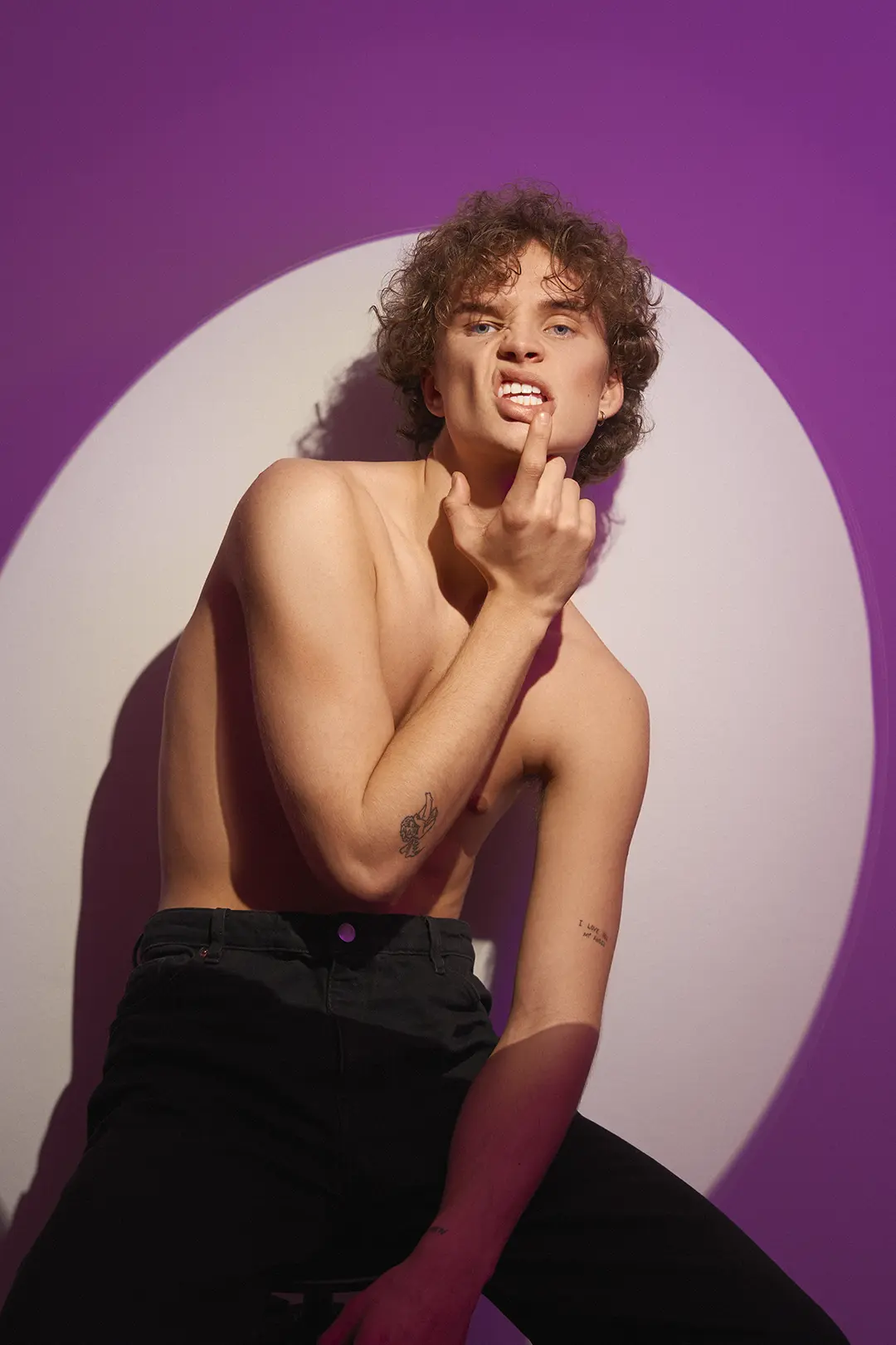 Ung shirtless person med lockigt hår och tatueringar poserar mot en lila och vit cirkelbakgrund.