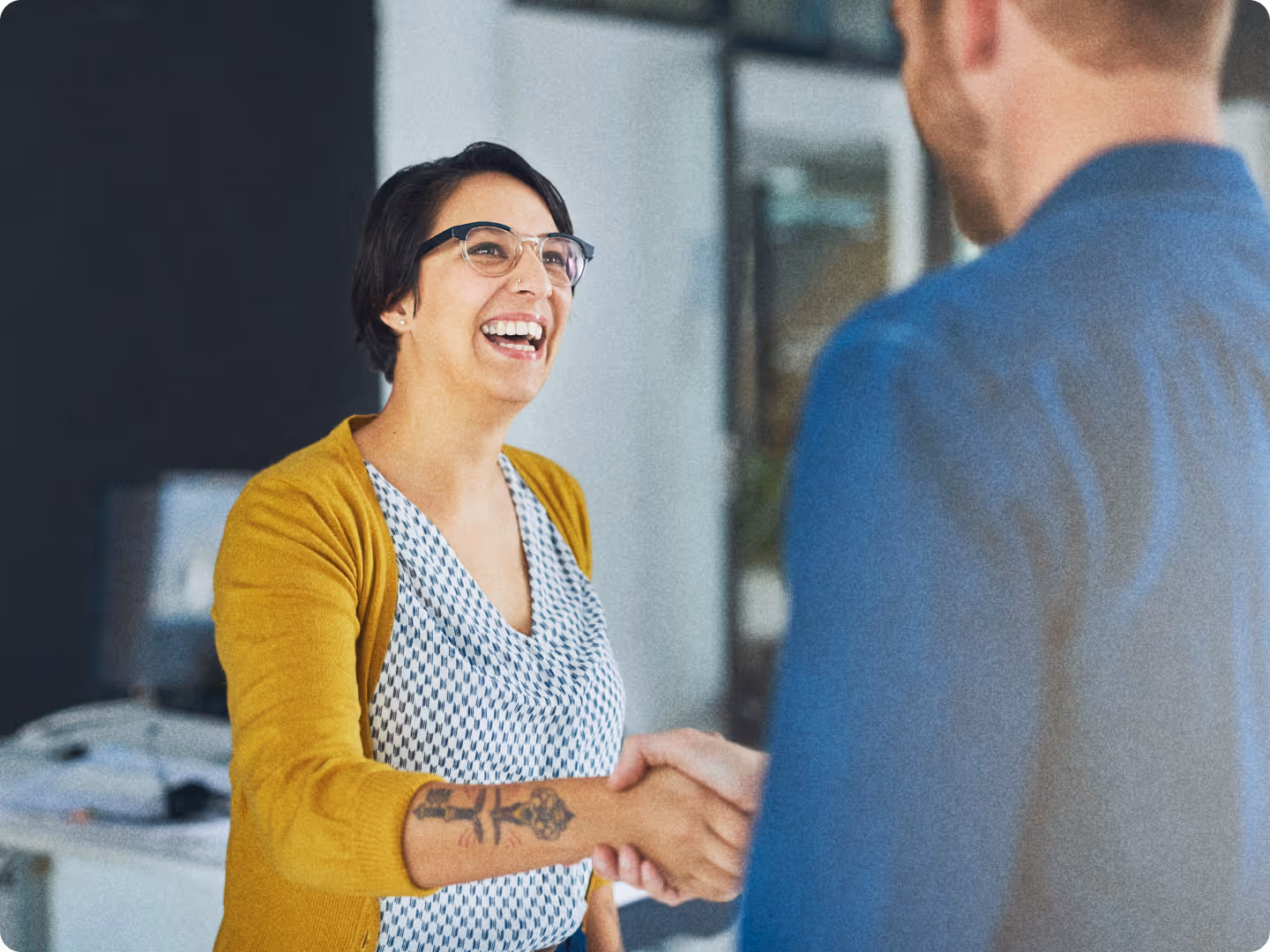 Two people shaking hands, one woman smiling and wearing glasses and a yellow cardigan.
