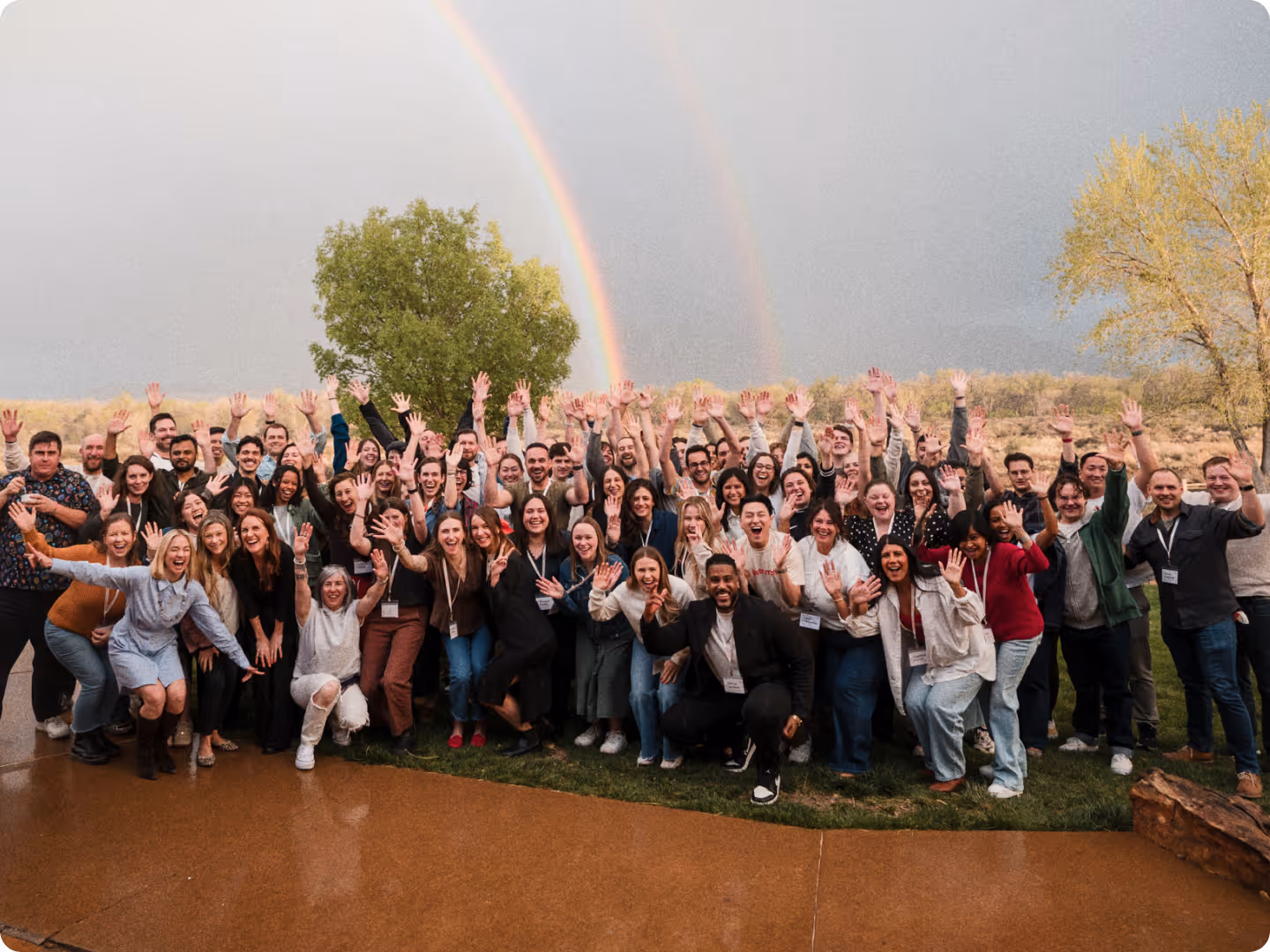 Large group of diverse people outdoors smiling and waving under a double rainbow.