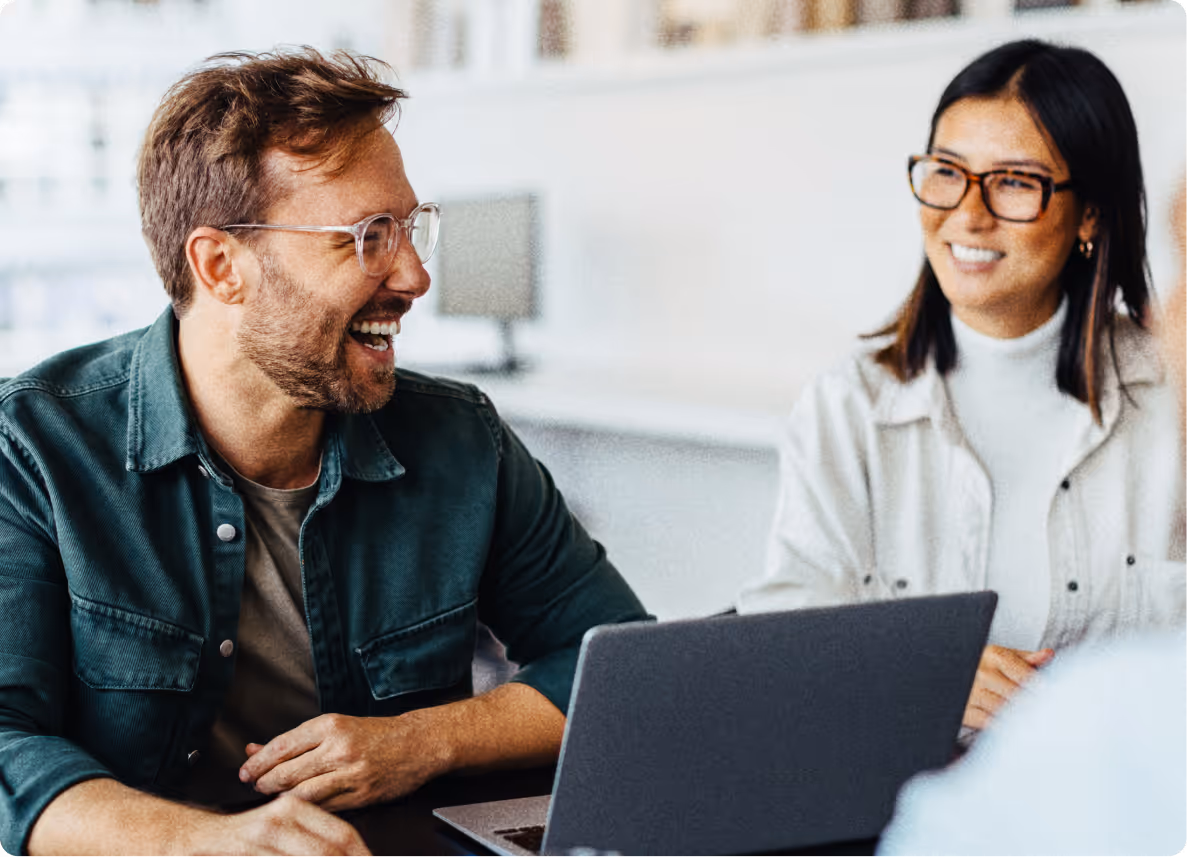 Man and woman with glasses sitting at a table, smiling and talking, with a laptop in front of them.