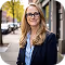 Smiling woman with glasses and long hair standing outdoors on a city sidewalk.