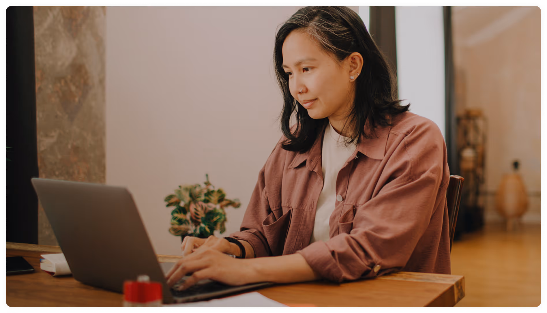 Woman wearing a mauve shirt typing on a laptop at a wooden table in a cozy room.