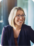Smiling woman with shoulder-length blonde hair and glasses wearing a dark blazer indoors.