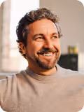 Smiling man with curly hair and beard, wearing a beige shirt, looking to the side indoors.