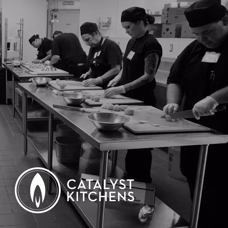 Five chefs in black uniforms preparing food on stainless steel tables in an industrial kitchen.